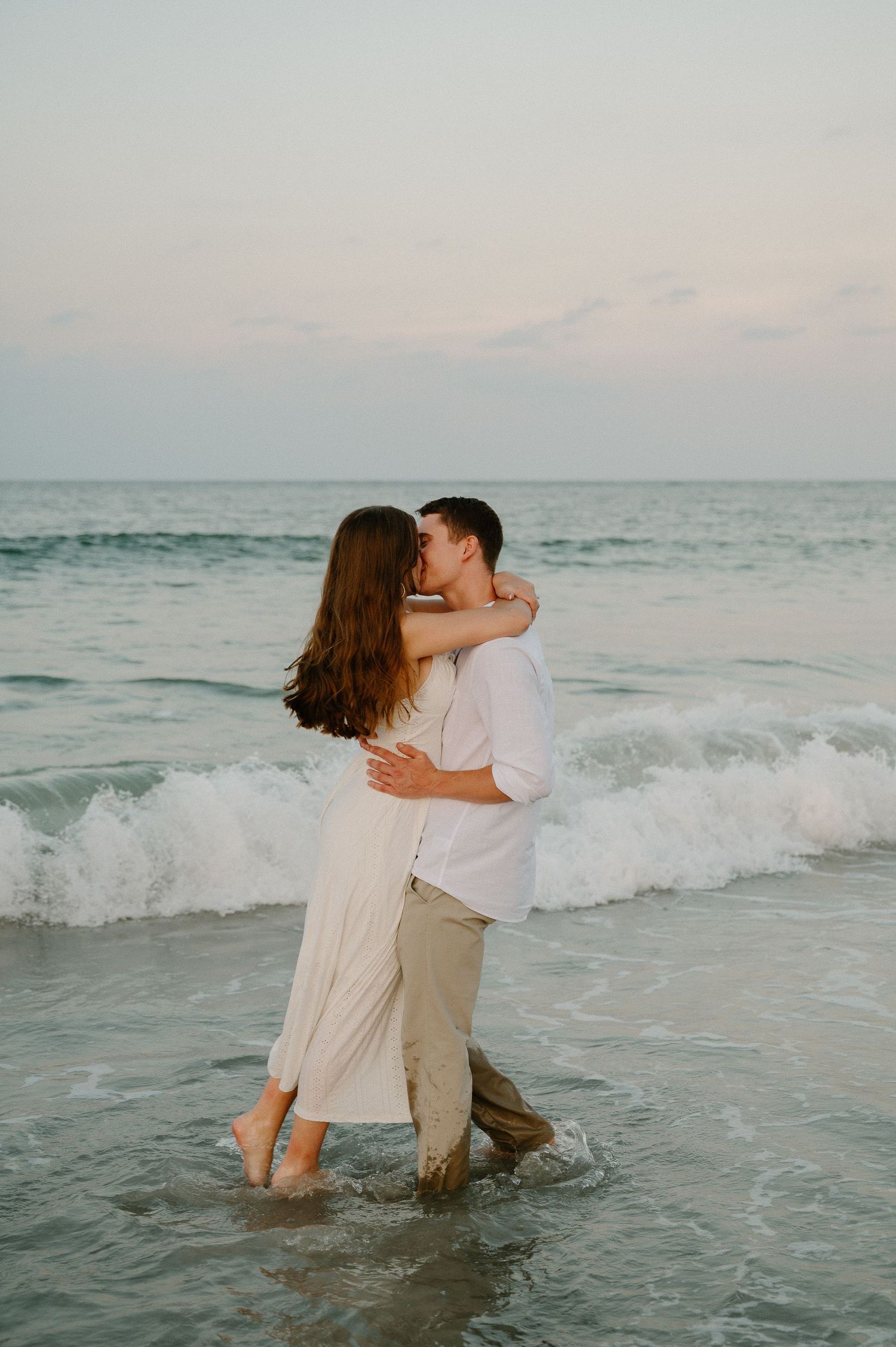 Couple kissing and embracing while standing in ocean waves at sunset on a sandy beach.