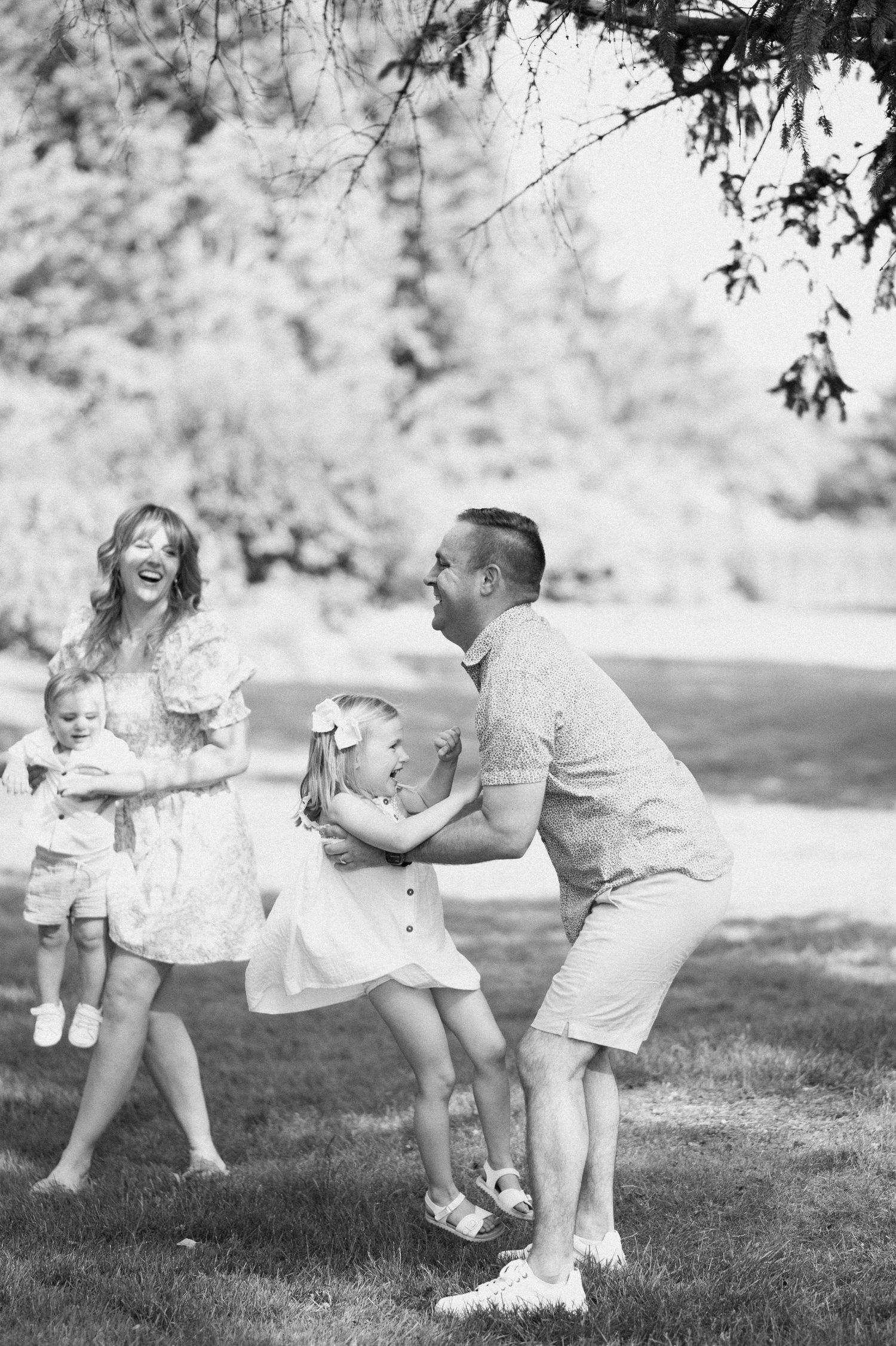 Family playfully interacting on grass in a sunlit outdoor setting wearing coordinated white and beige outfits.