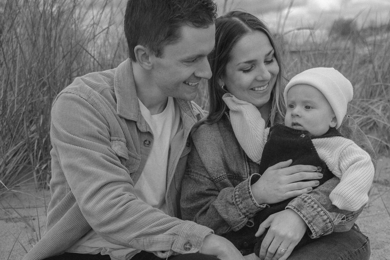 Black and white photo of a smiling couple sitting on a beach with their baby in a white hat.