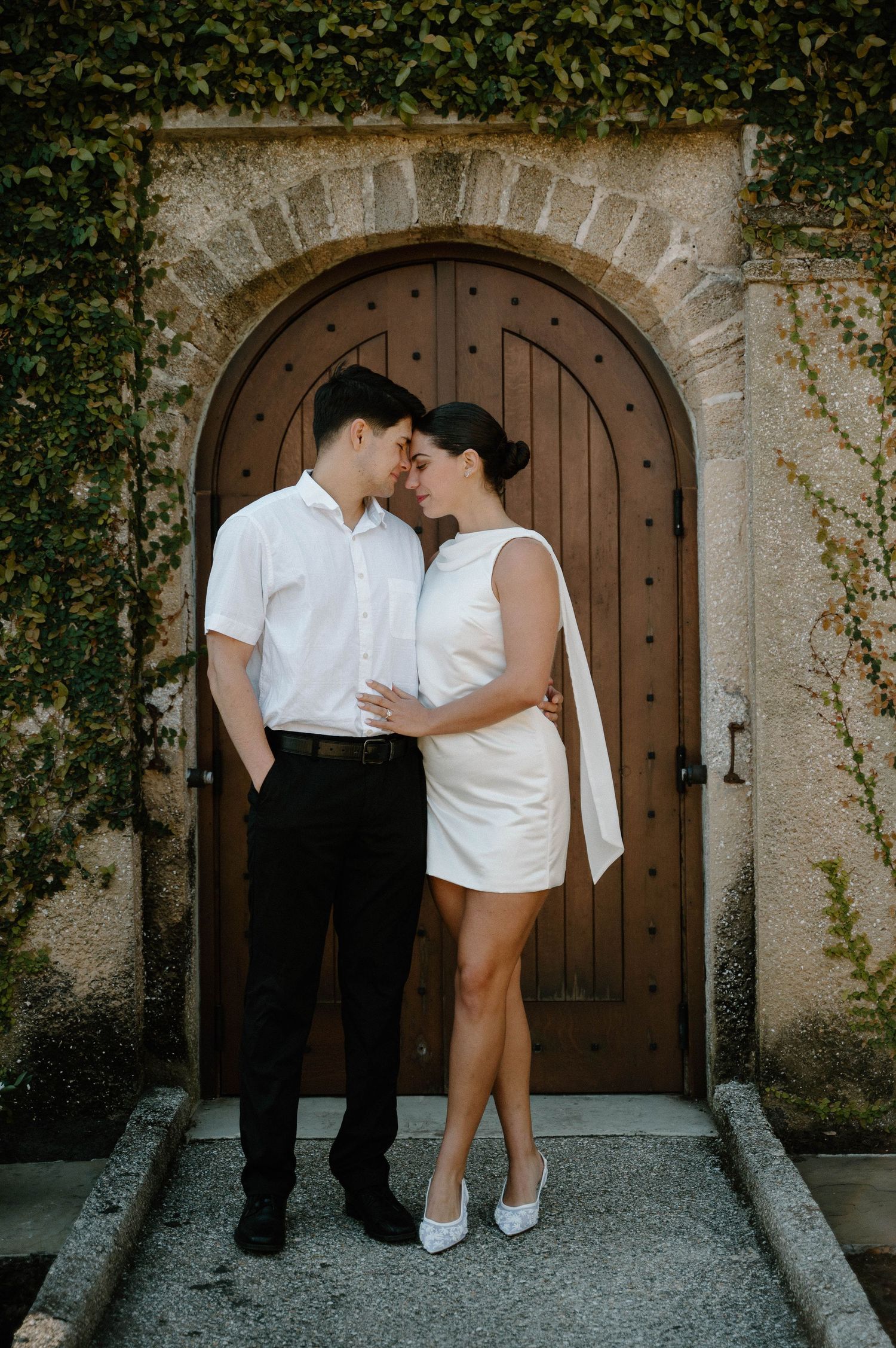 Couple in white outfits sharing a romantic moment in front of an arched wooden door covered in ivy.