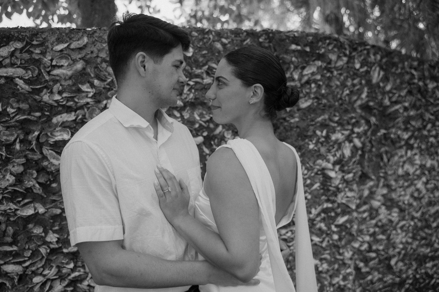Black and white photo of a couple in wedding attire sharing a romantic moment outdoors near a leafy wall.