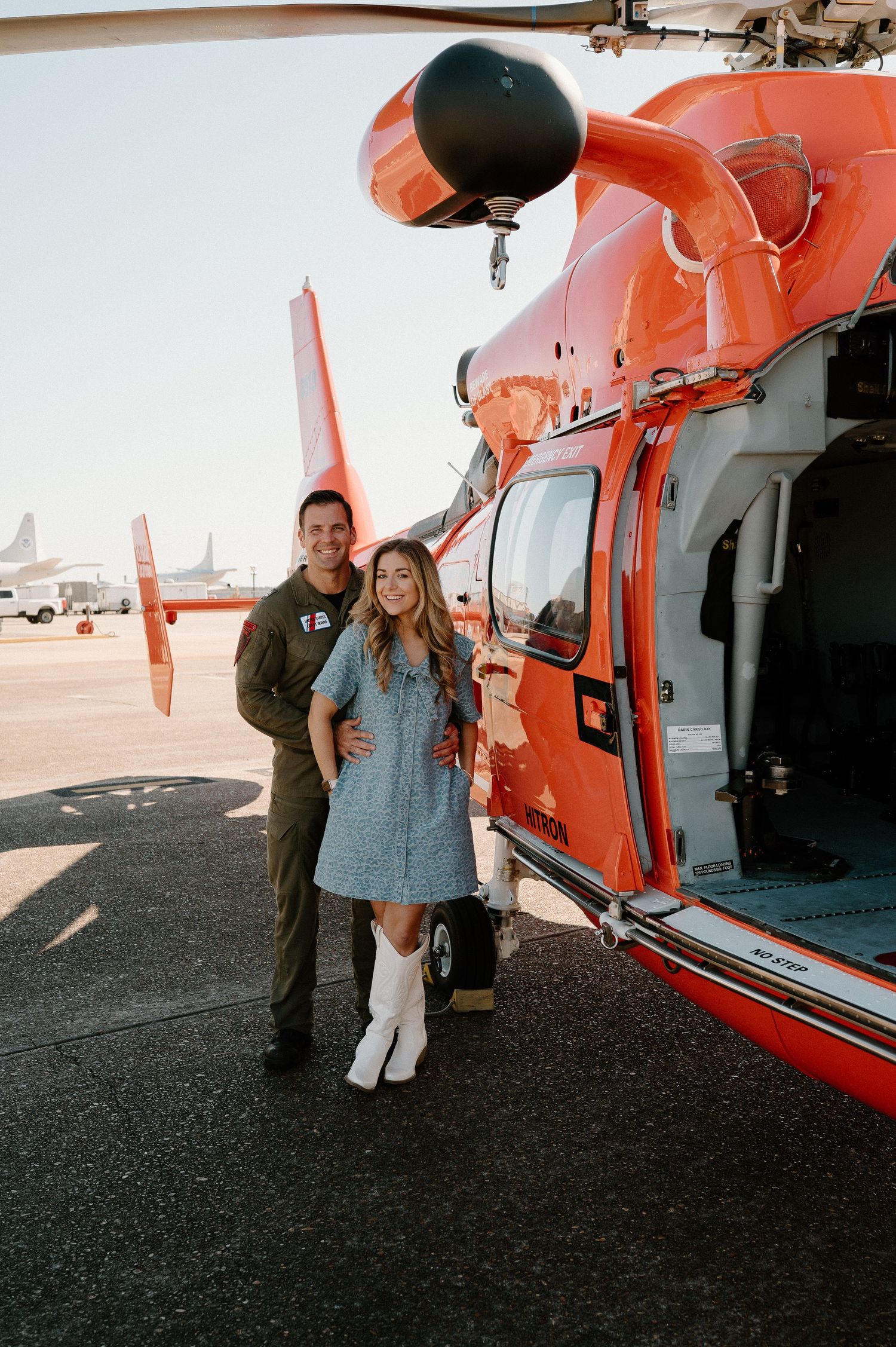 Couple posing beside an orange US Coast Guard helicopter on a sunny airfield tarmac.