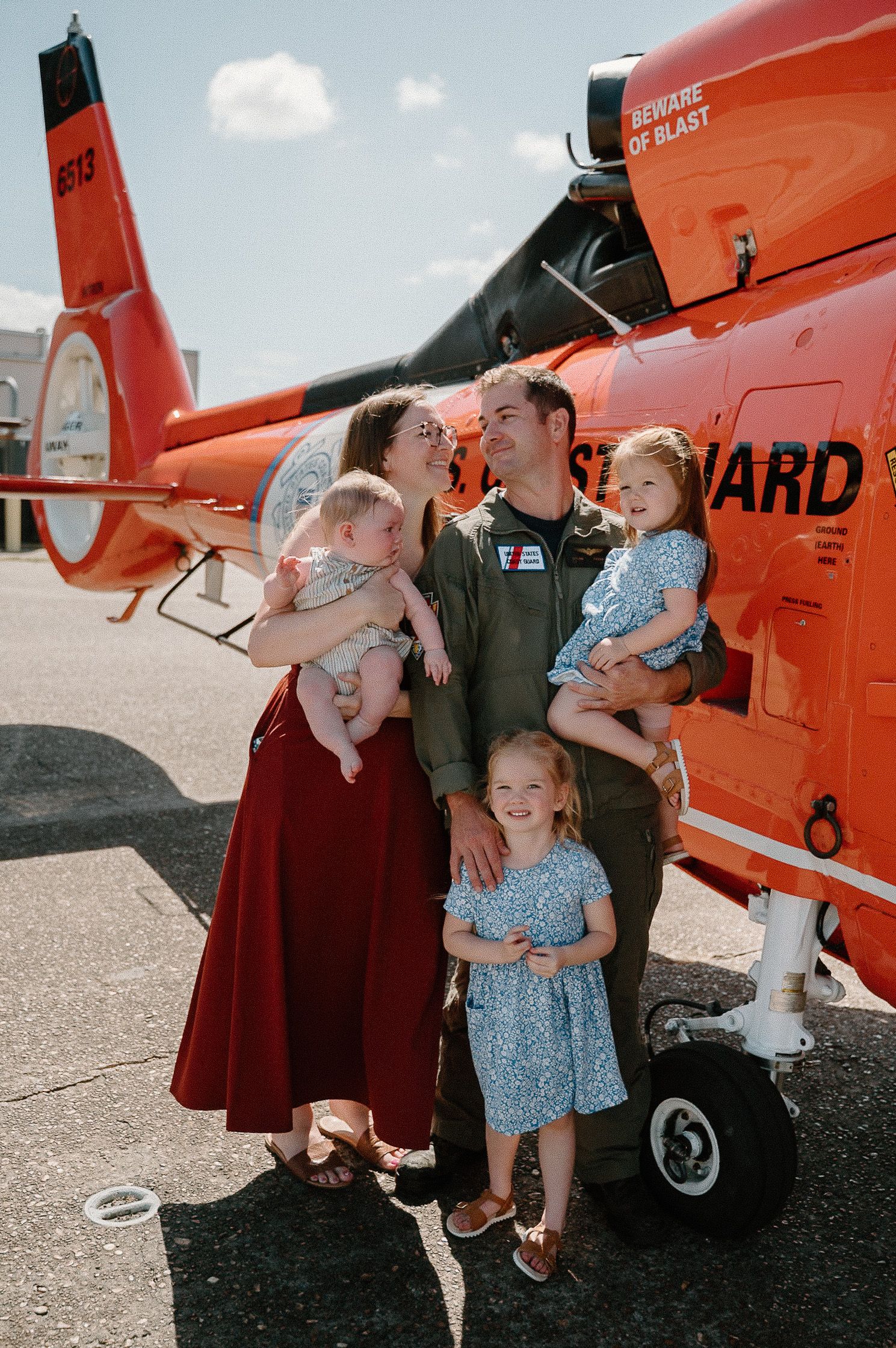 Coast Guard pilot poses with family in front of orange helicopter on sunny tarmac, holding children in festive attire.