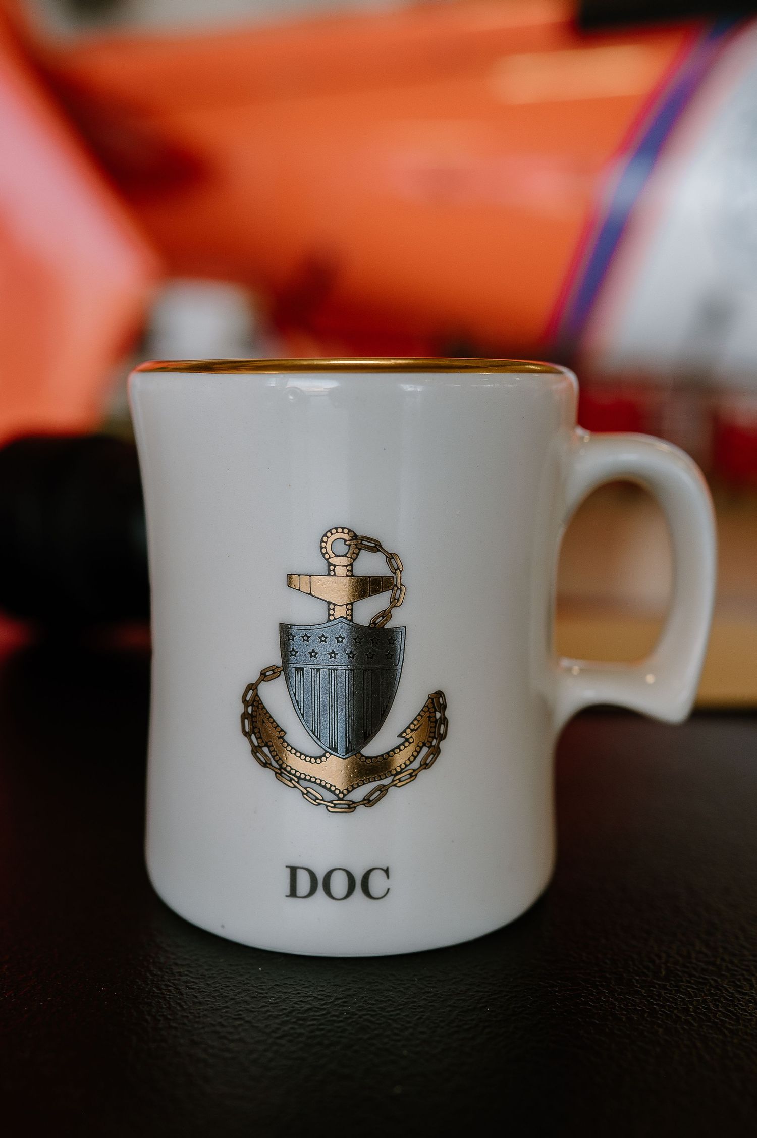 White ceramic mug with gold rim featuring a nautical anchor emblem and the text DOC on a dark surface.