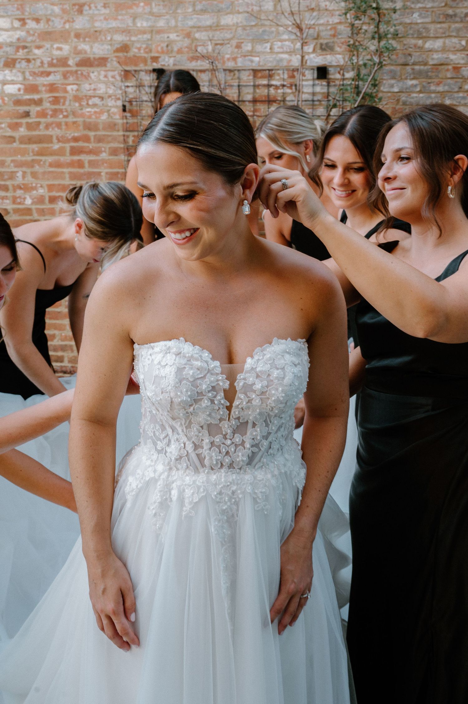 Bride in floral strapless wedding gown smiles as bridesmaids in black dresses help her get ready near brick wall.