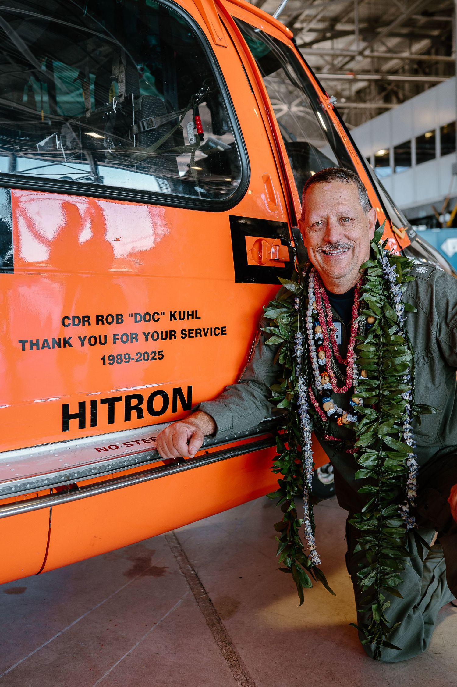 CDR Rob Doc Kuhl poses beside an orange Hitron helicopter honoring his 1989-2025 military service, wearing Hawaiian leis.