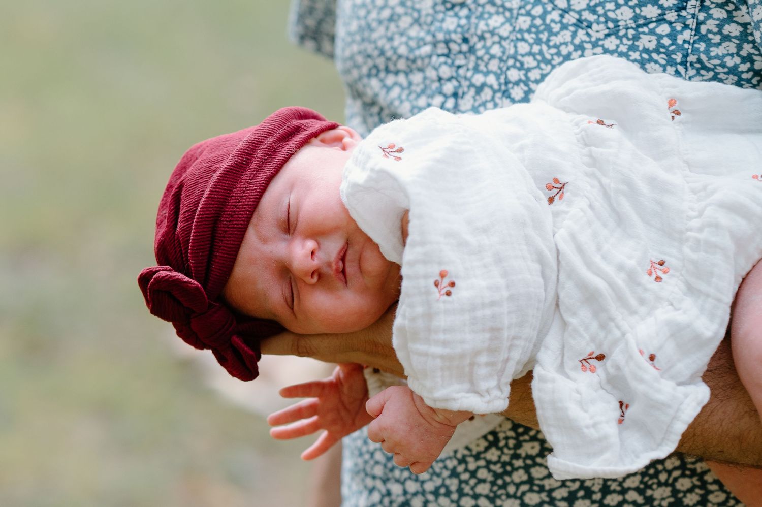 A small baby wearing a burgundy hat and white dress is held close.
