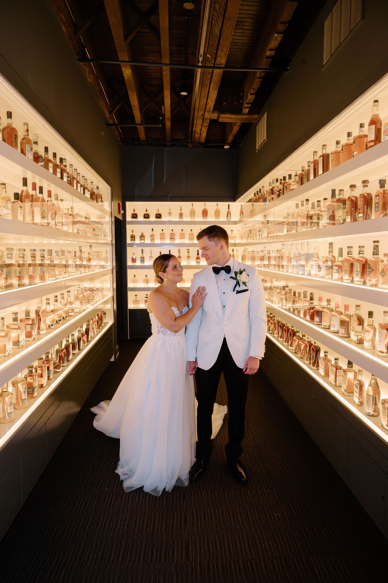A couple poses dramatically in an illuminated room with walls of bottled spirits.
