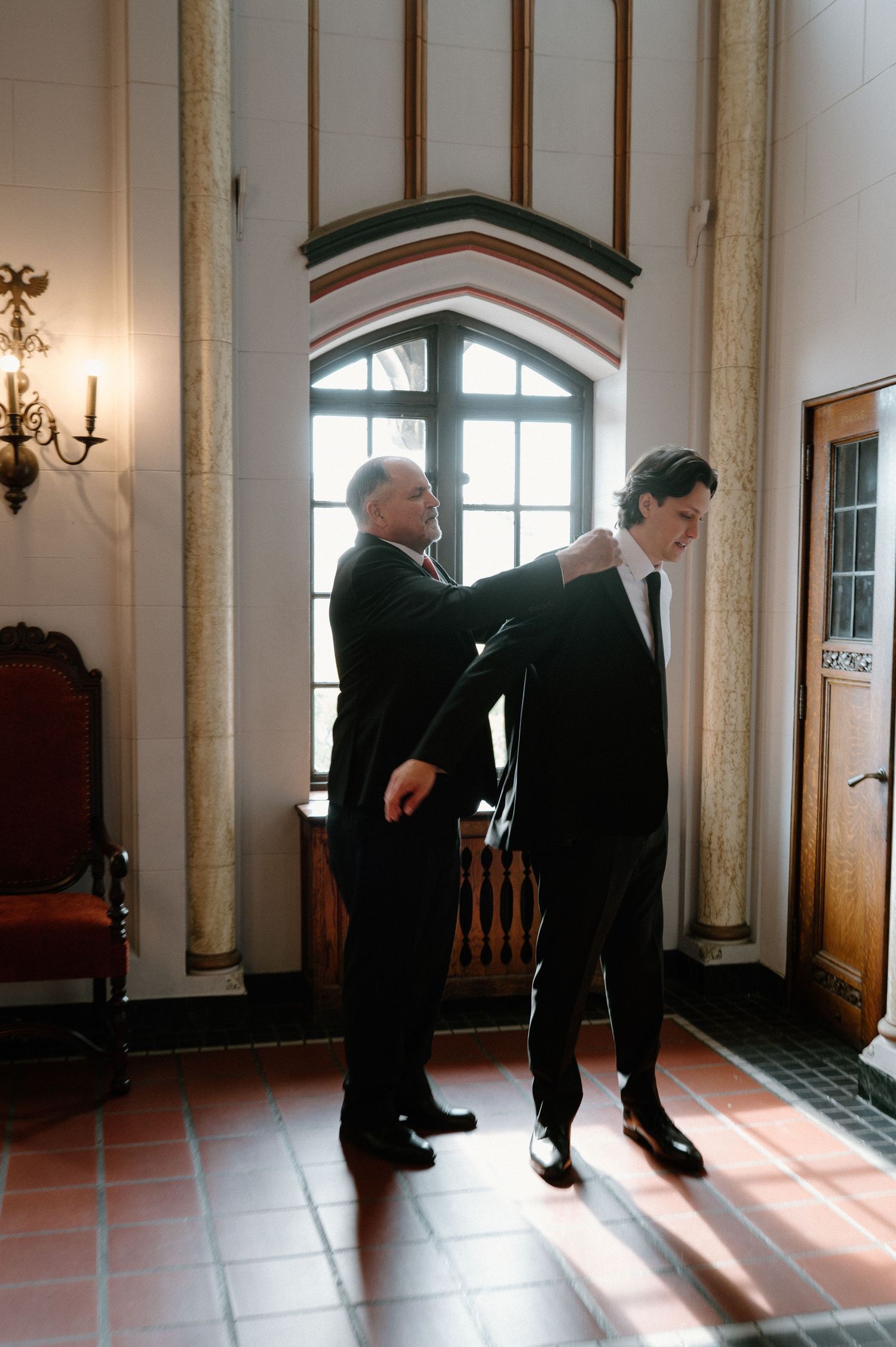 Two men in formal attire stand in an elegant hallway with arched windows and wall sconces.