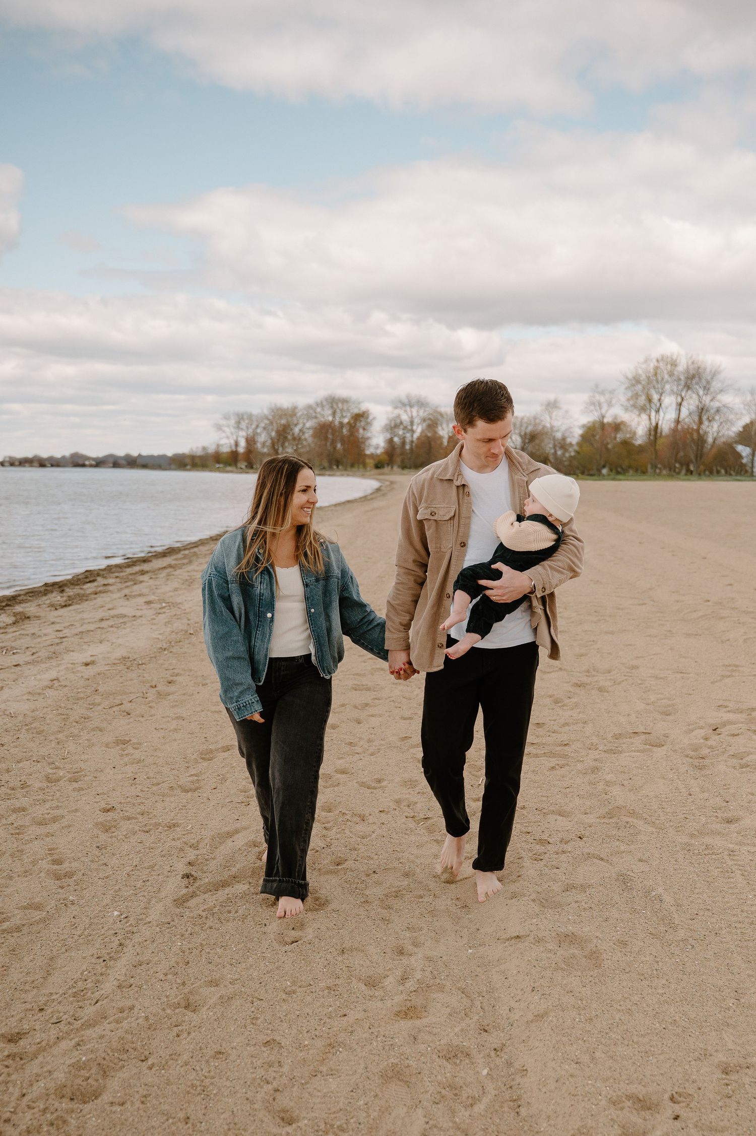 A young couple walks hand-in-hand on a sandy beach, with the man cradling a newborn baby.