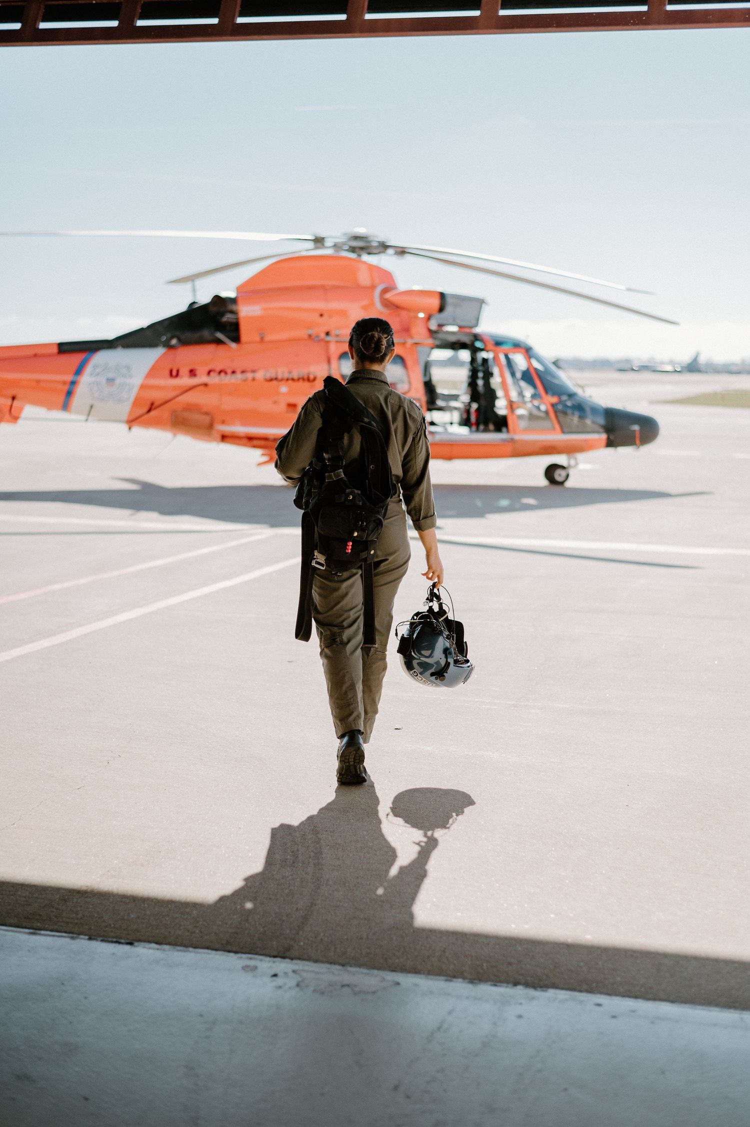 Coast Guard pilot walking towards orange helicopter carrying flight gear.