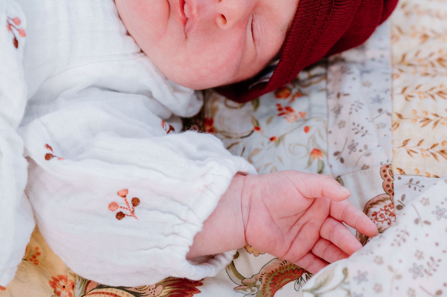 A sleeping newborn in a red cap and white outfit rests on a patterned blanket.
