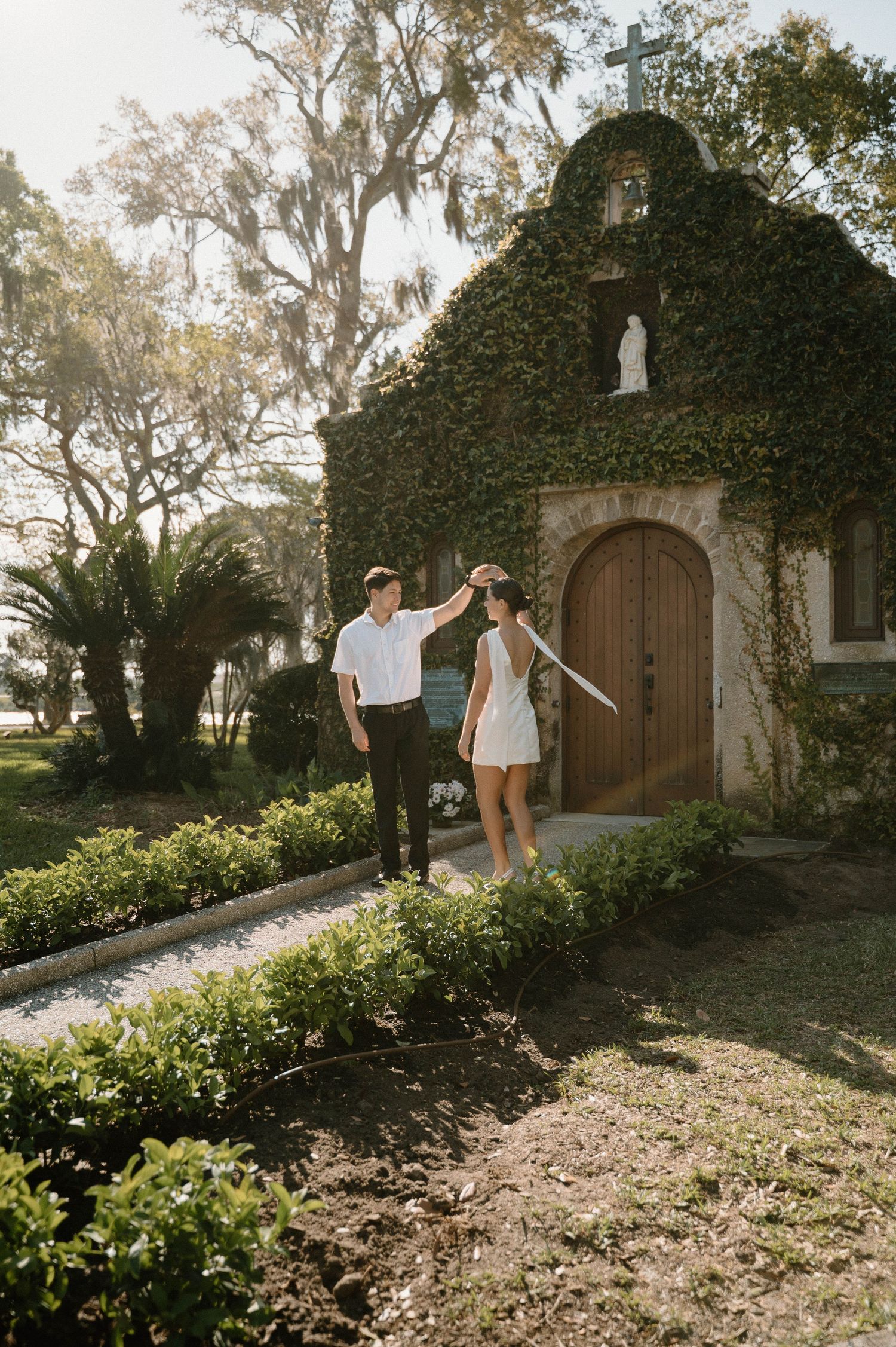 Couple dancing near ivy-covered chapel with wooden arched door and statue, surrounded by lush greenery in warm sunlight.