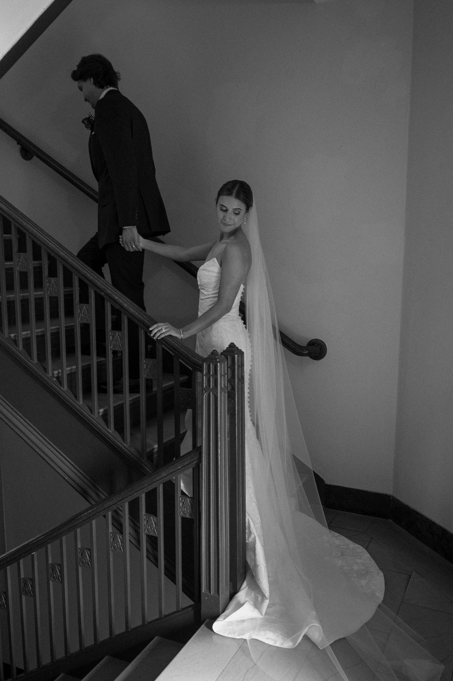 A bride in a white gown and veil poses on a staircase while a figure in black ascends the stairs in black and white photography.