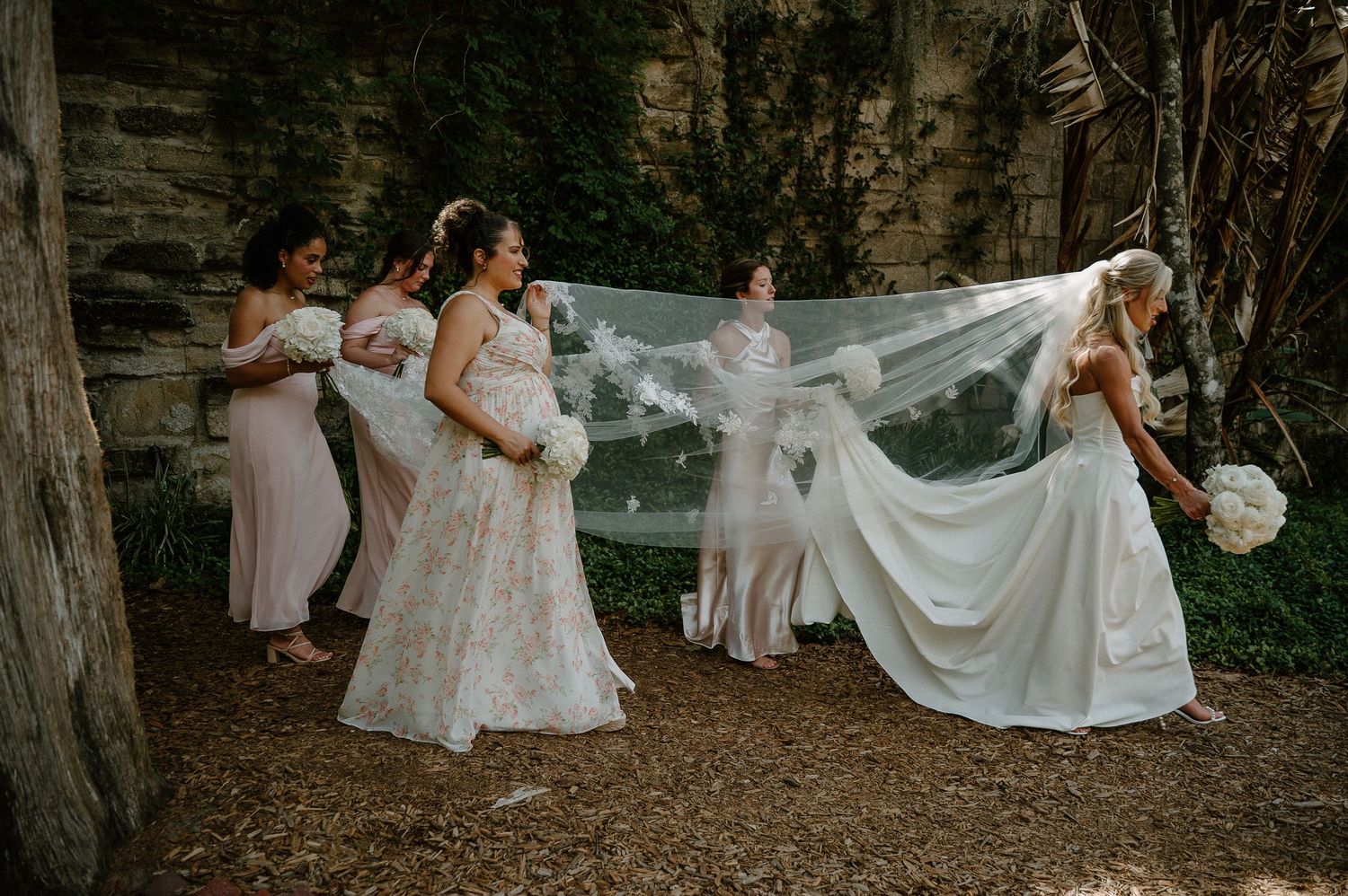 Bride in floral gown with bridesmaids in pink satin dresses holding long veil outdoors near stone wall.