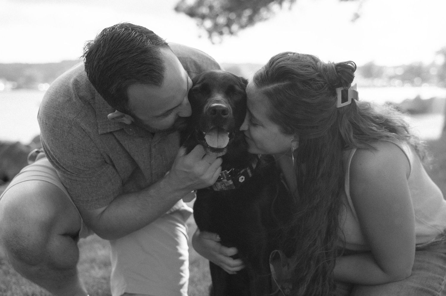 Black and white photo of a couple kissing their happy black Labrador dog outdoors near a lake.