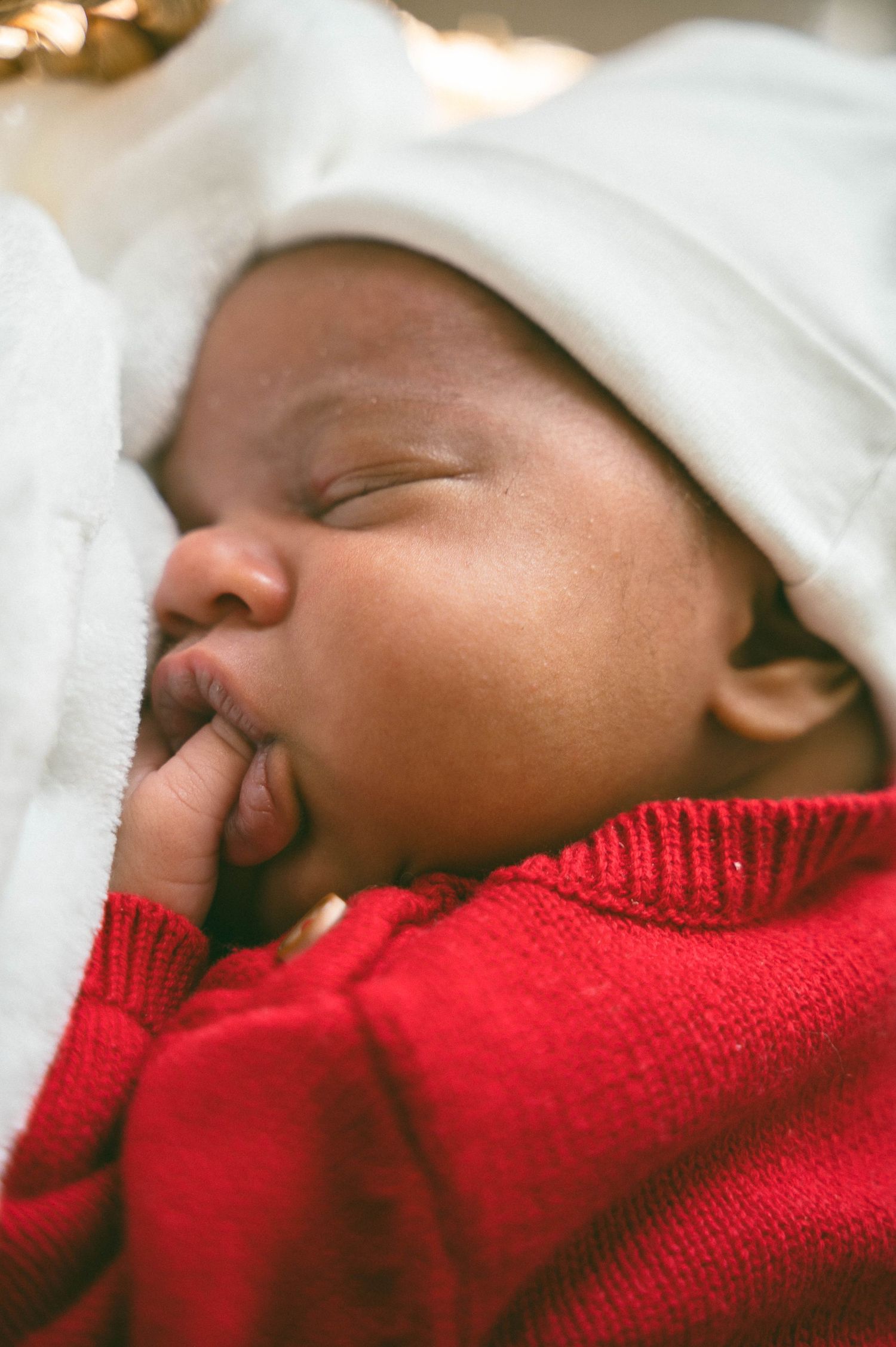 Peaceful newborn baby sleeping in a white hat and red knit sweater, cradled in soft white blankets.