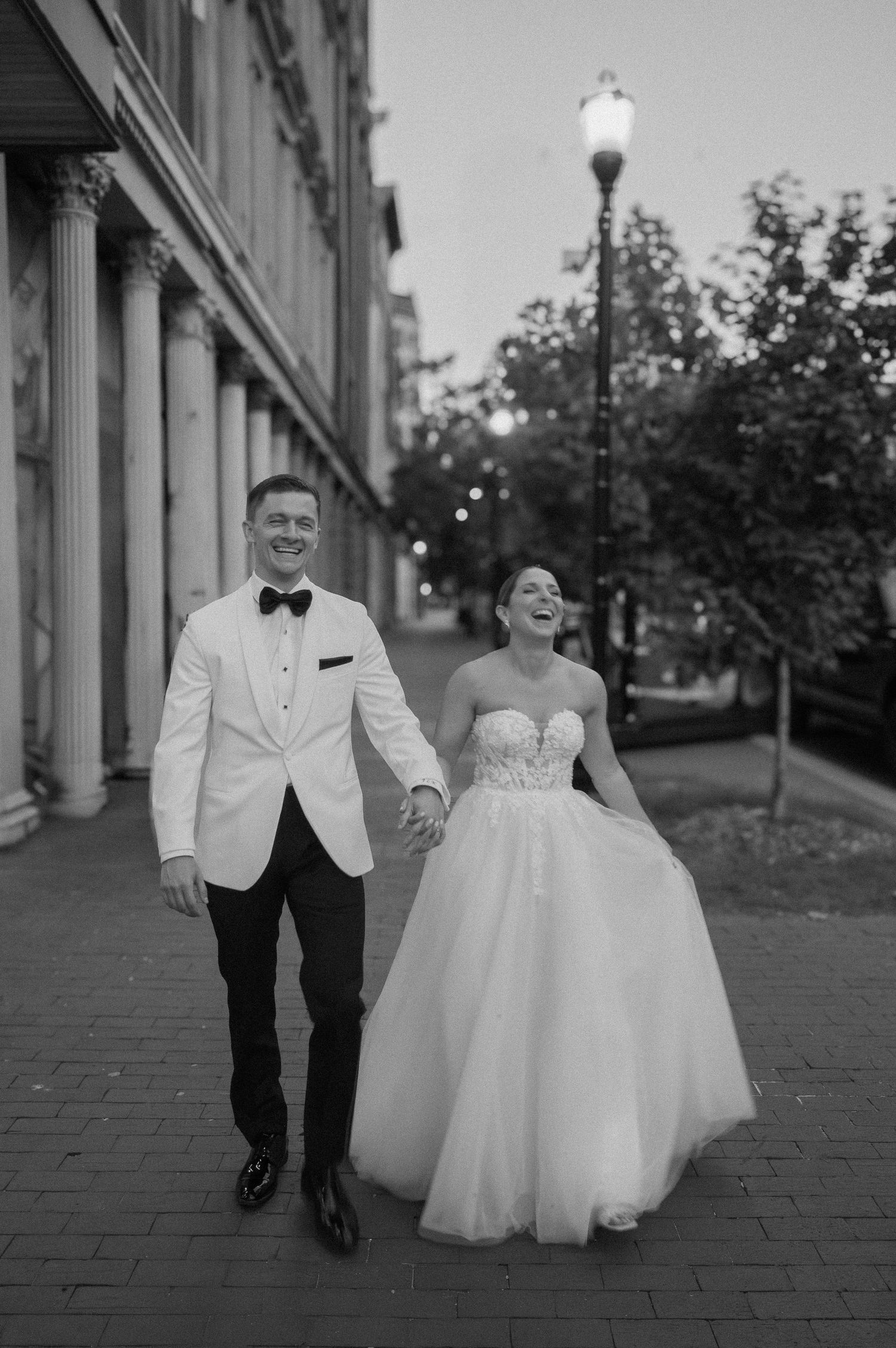Couple walks along brick sidewalk in formal wedding attire.