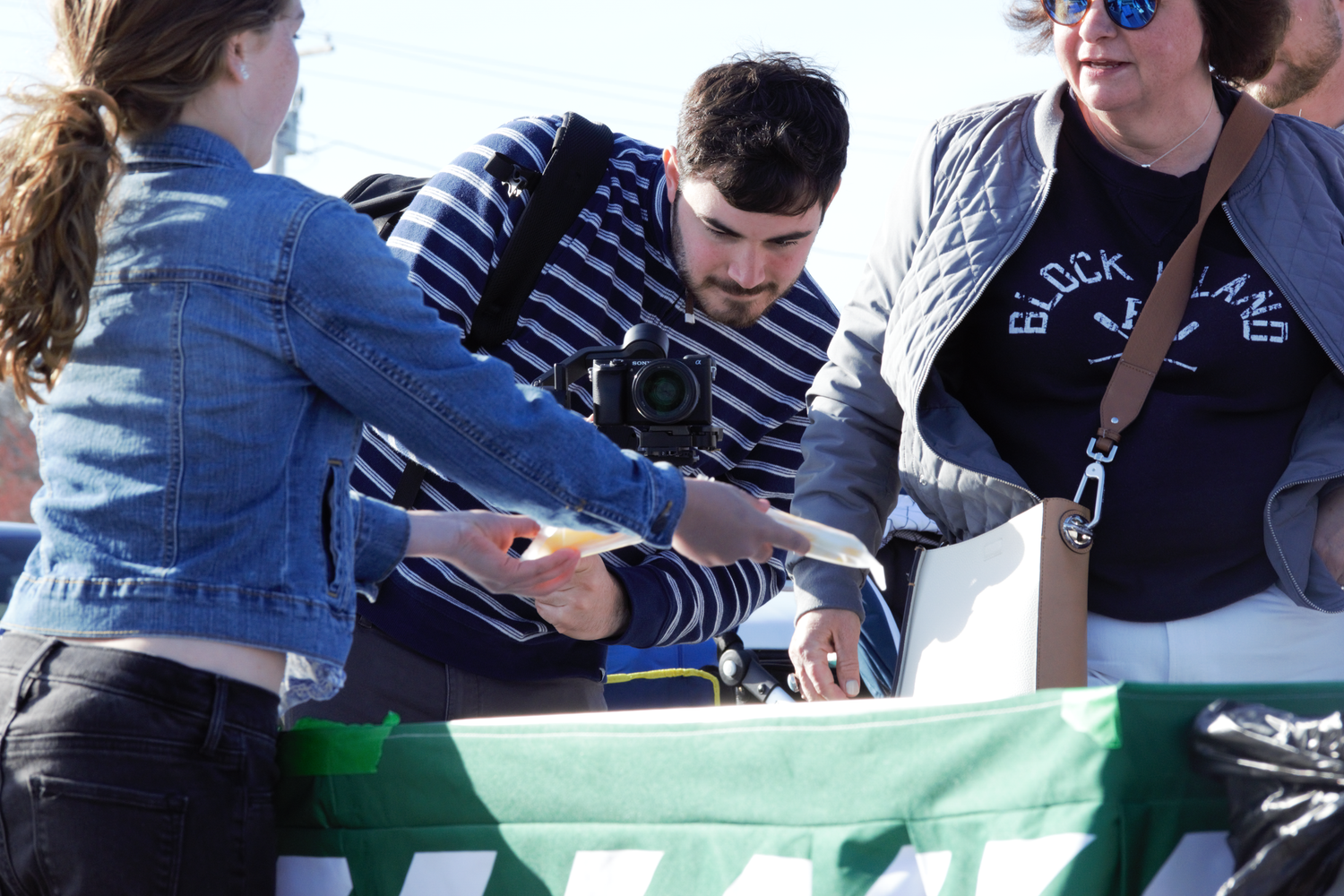 People gathered outdoors at a table, with a man in a striped shirt using a camera at a sunny event.