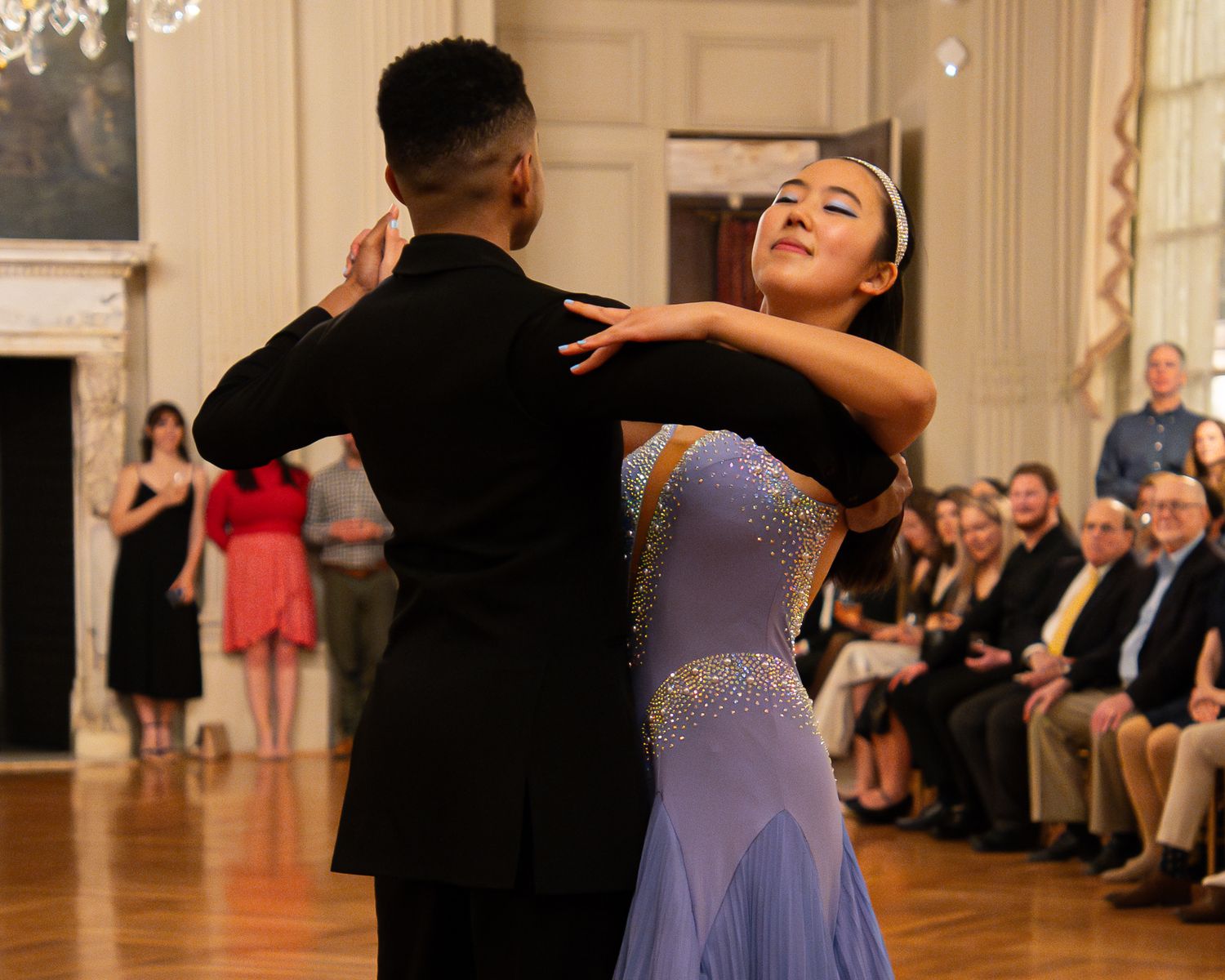 Couple performing an elegant ballroom dance with the woman wearing a purple dress and man in black.