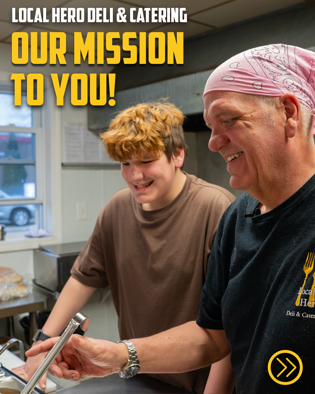 Two smiling deli workers at Local Hero Deli & Catering preparing food together behind the counter.