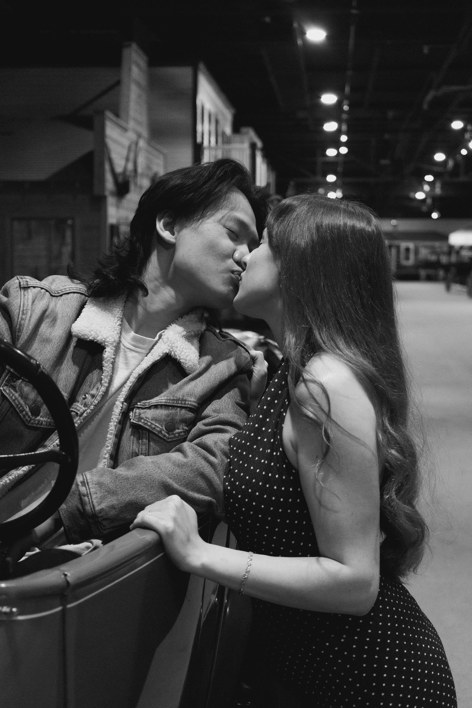 Black and white photo of a couple sharing a romantic kiss near a vintage car in a dimly lit venue.
