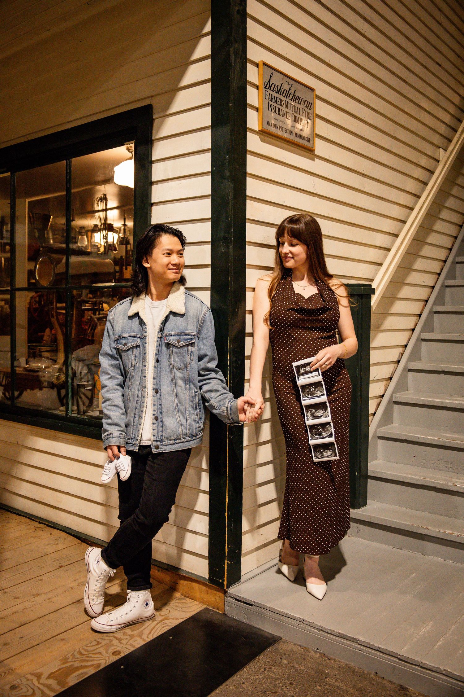 Couple holding hands outside vintage shop, woman in polka dot dress holding ultrasound photos, announcing pregnancy.