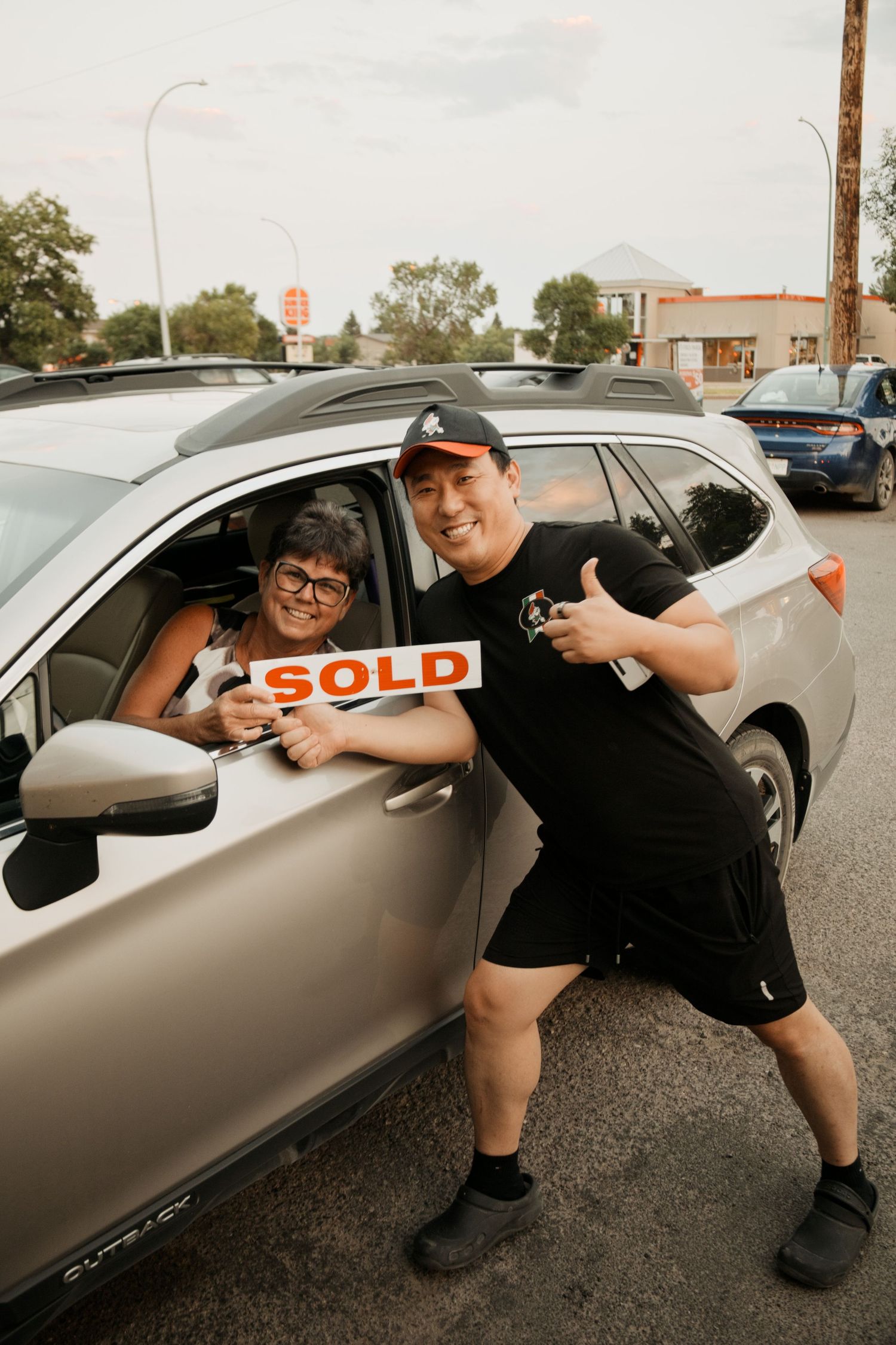 Two people celebrating a car sale, holding a red SOLD sign beside a silver SUV in a parking lot.