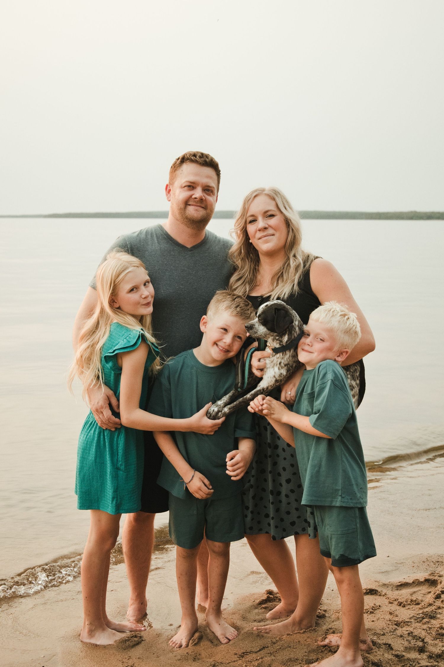 Happy family of two adults and three children posing with a spotted dog on a sandy beach near calm water.