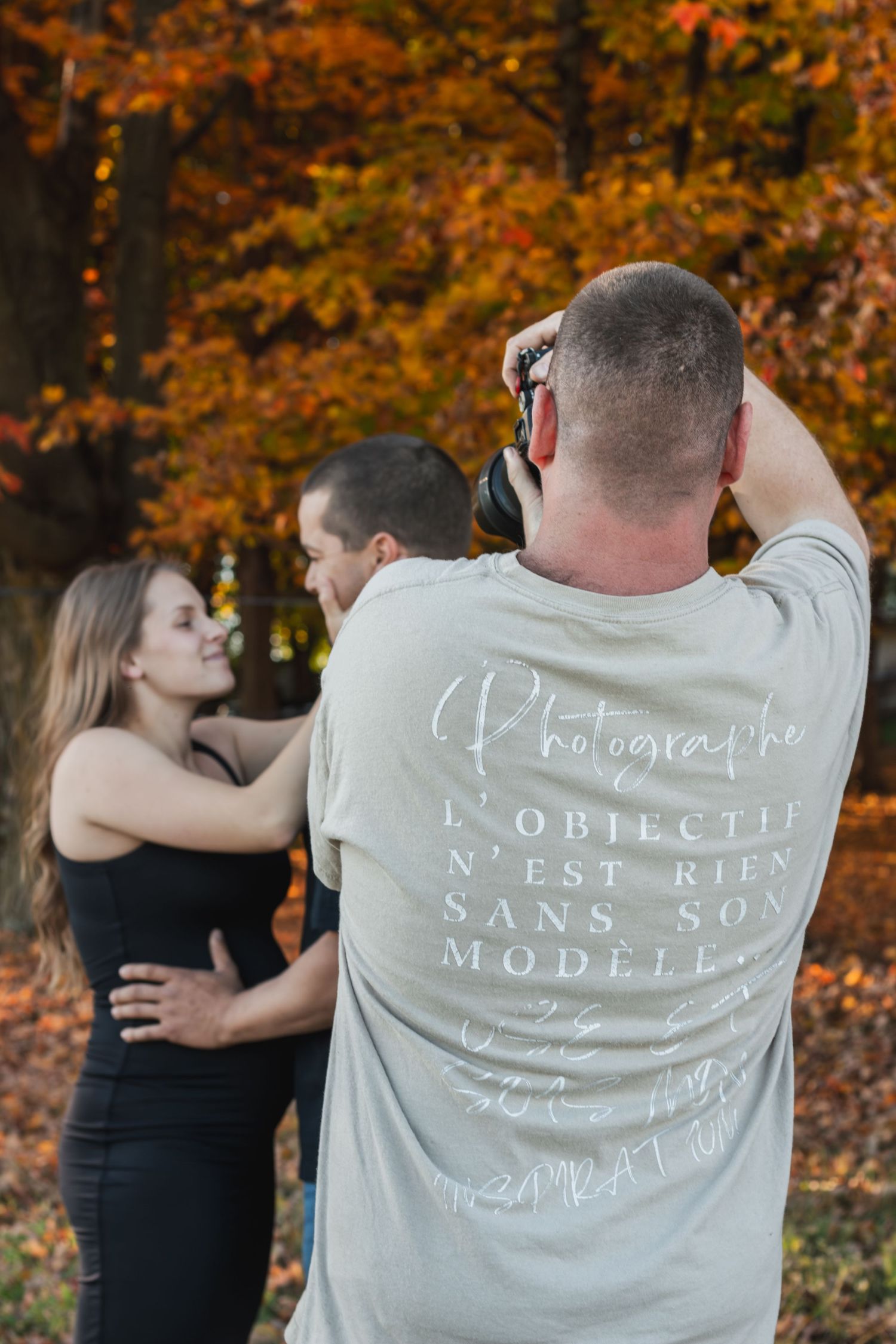 A couple poses together in an autumn setting with orange fall foliage in the background.