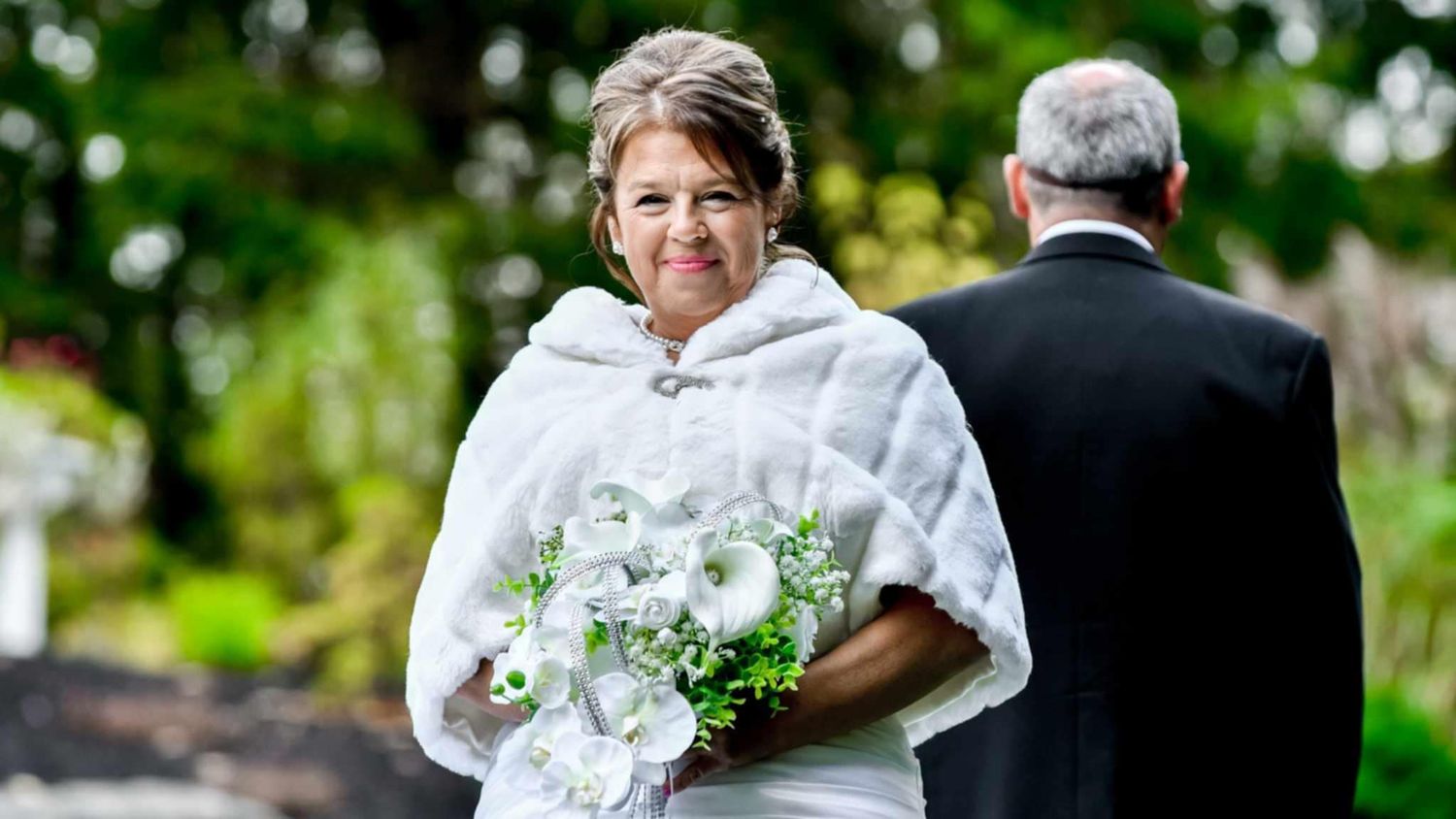 A wedding couple walks together through a lush garden setting with white floral bouquet and formal attire.