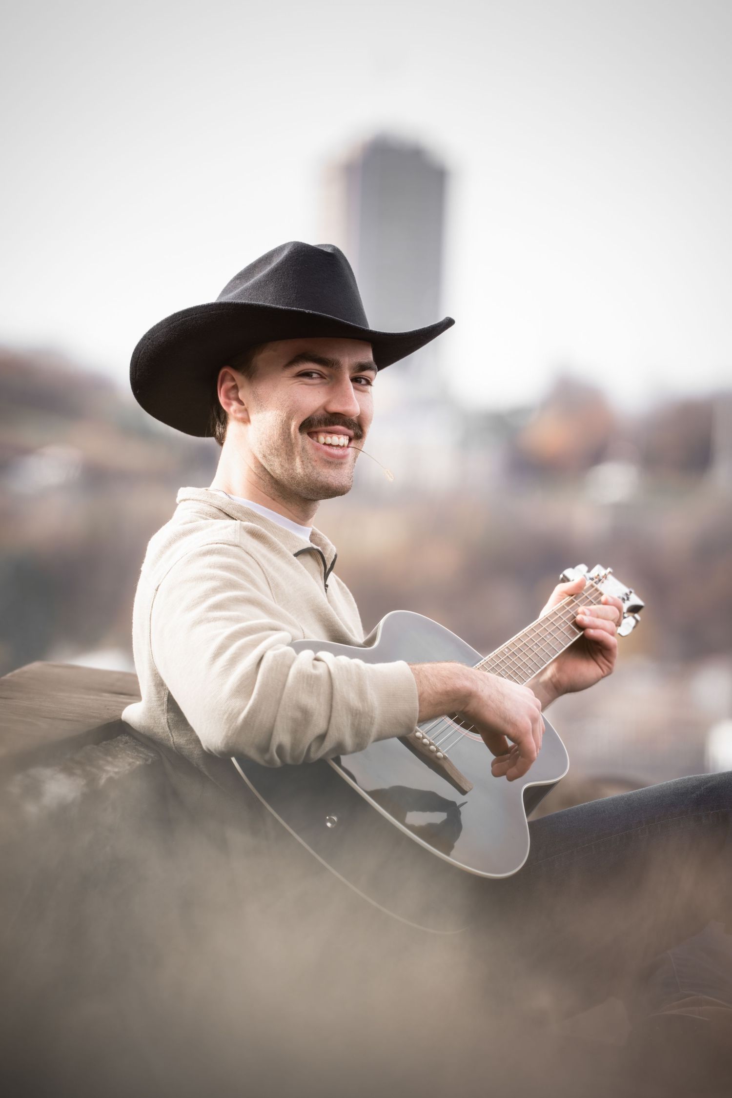 Musician in cowboy hat plays acoustic guitar outdoors with city skyline in background.
