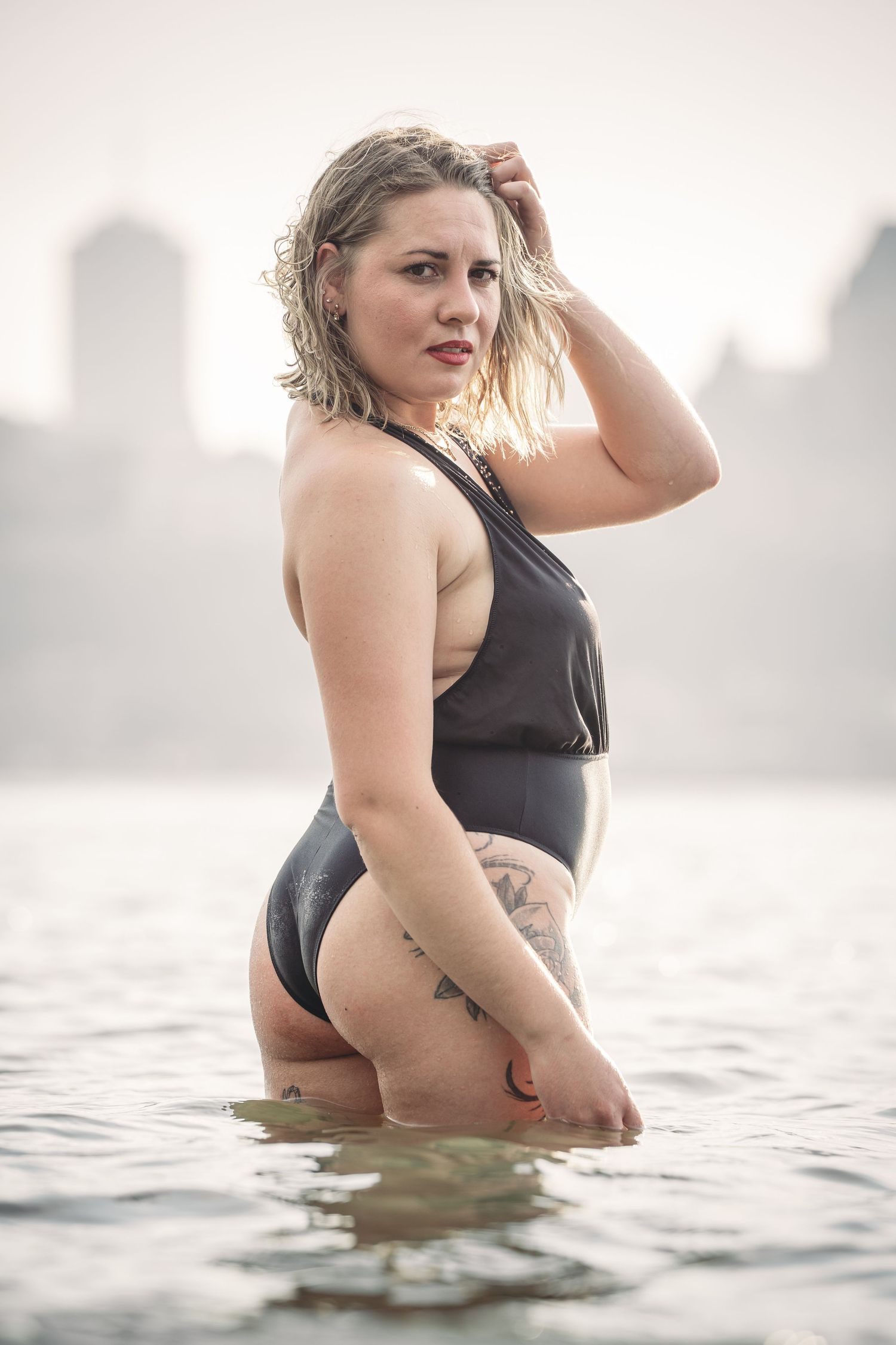 Woman in black swimwear poses gracefully in water with city skyline in background.
