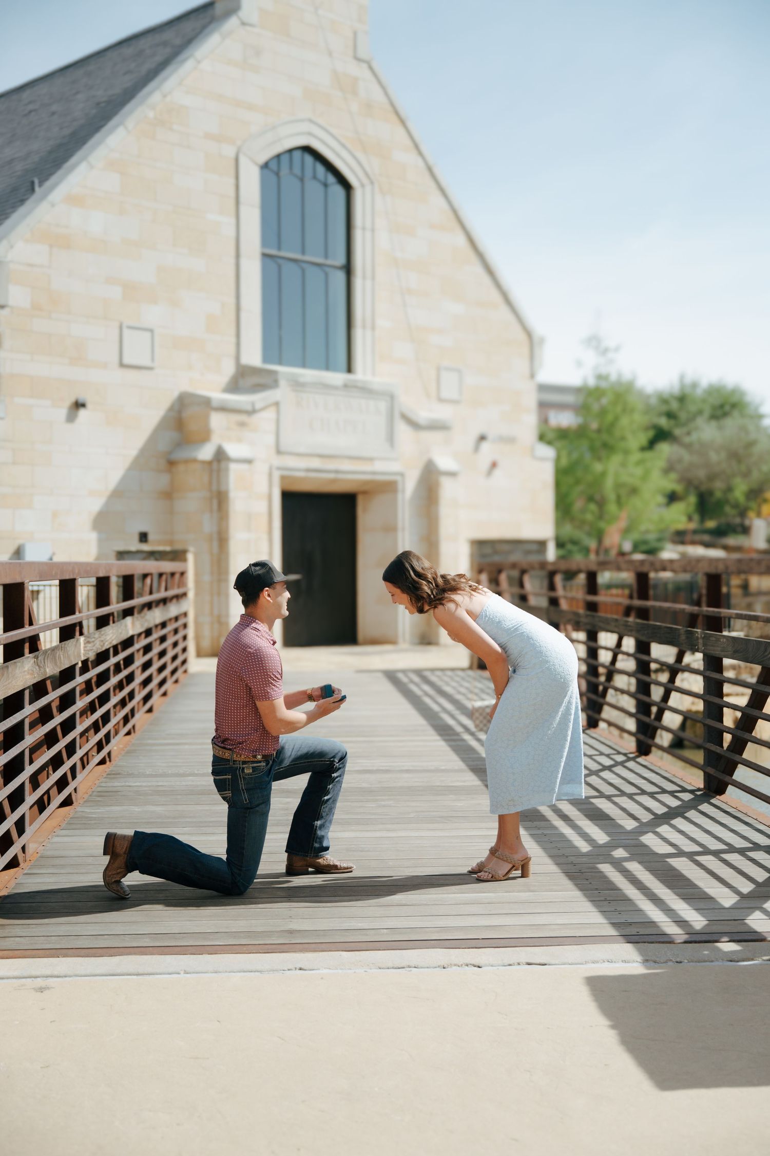 Man proposing on one knee to woman on wooden bridge outside stone church on sunny day.