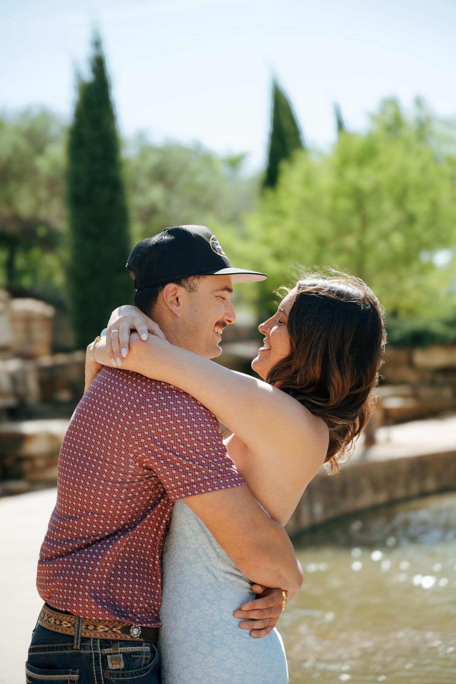 Happy couple embracing outdoors near a fountain, surrounded by lush green trees on a sunny day.