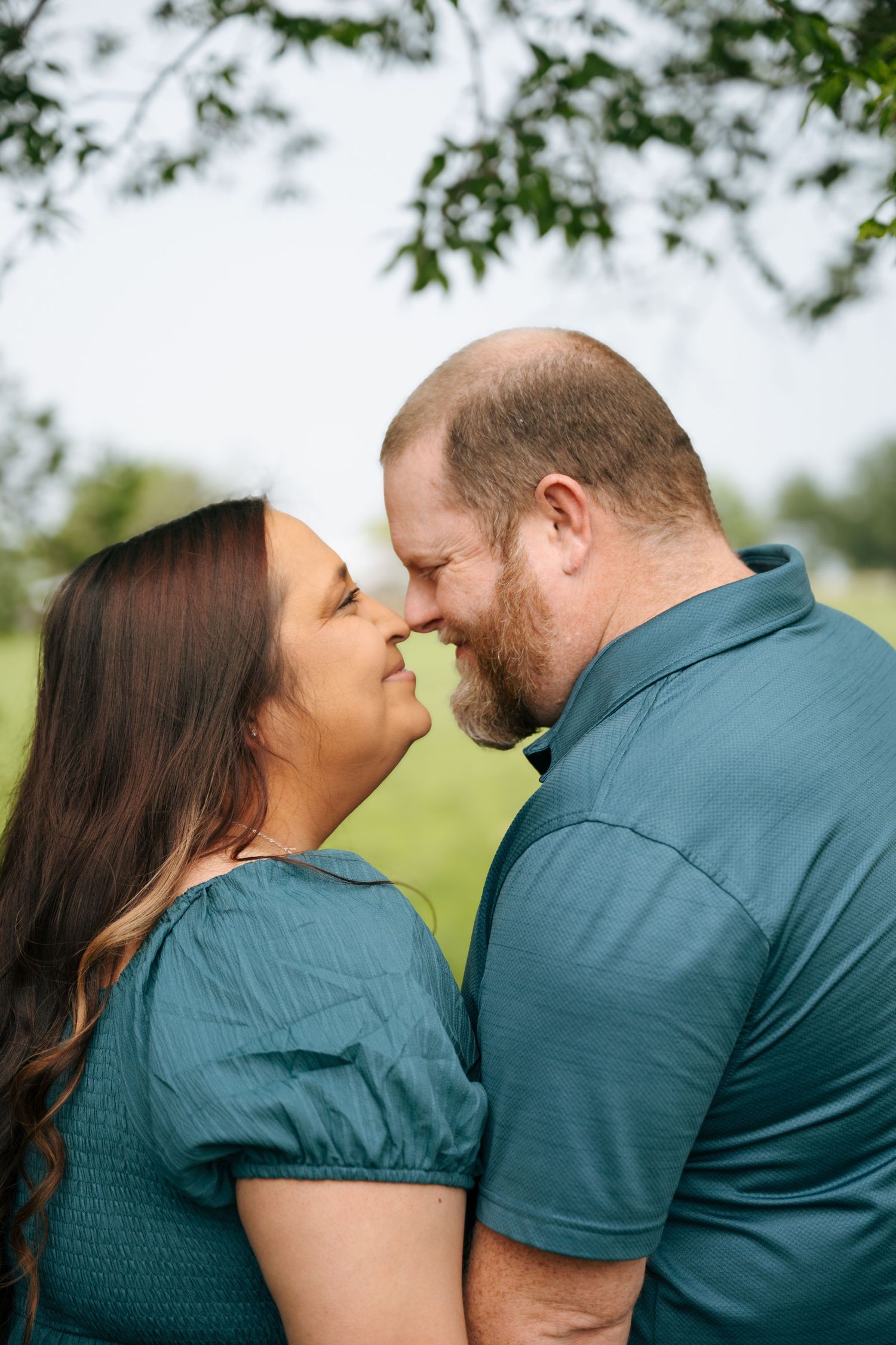 A couple in matching teal outfits share a tender nose-to-nose moment outdoors with green grass and trees behind them.