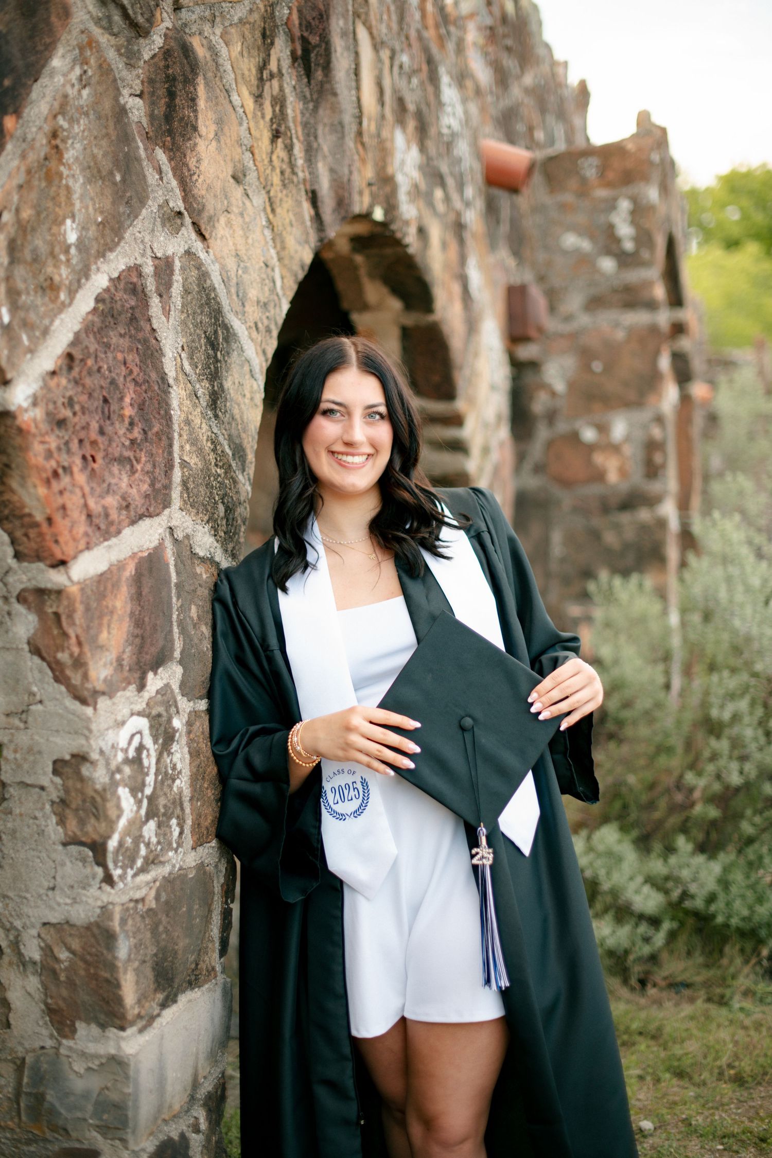 Graduate in dark green gown poses with diploma against stone wall.