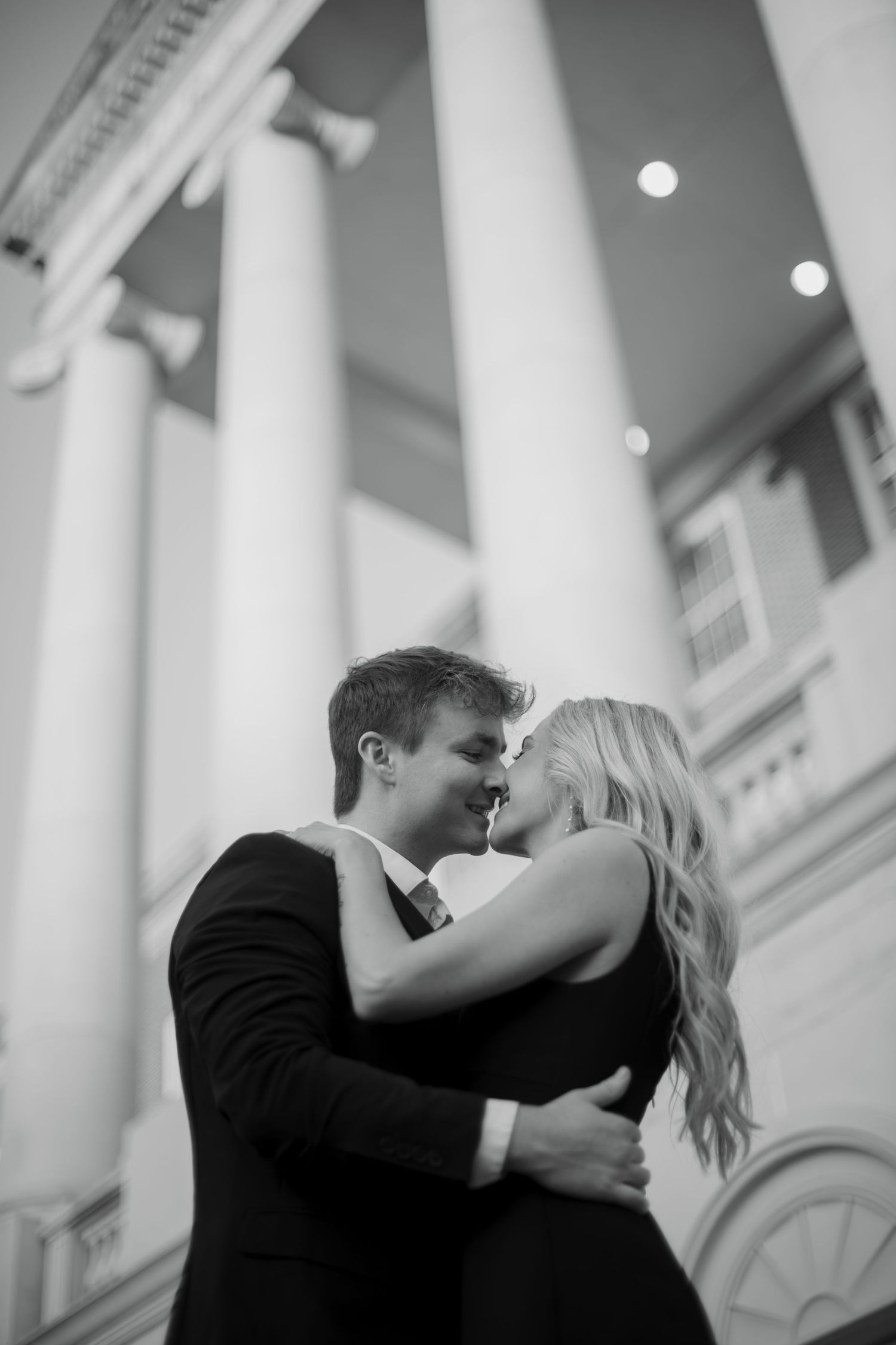 A romantic couple embraces and kisses in front of a classic white columned building in black and white photography.