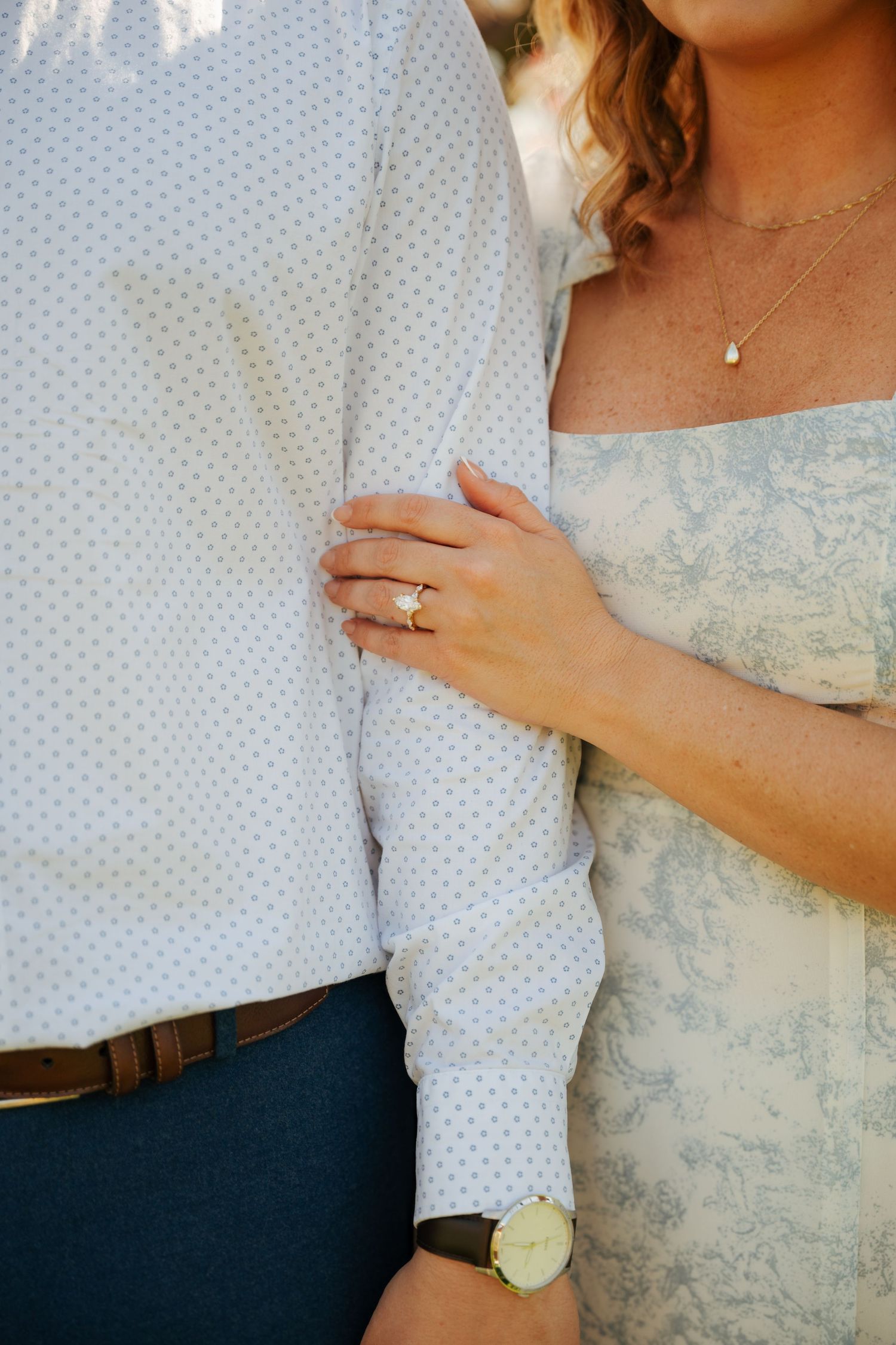 Close-up of engaged couple, woman wearing diamond ring and delicate necklace, holding mans arm outdoors.