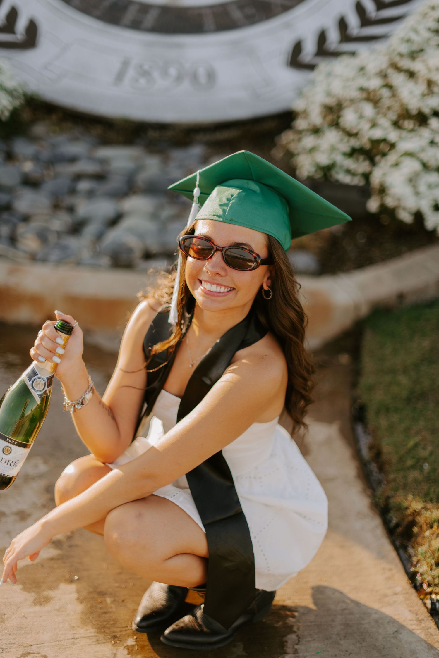 Graduate in green cap and white dress celebrates with champagne bottle on sunny day.