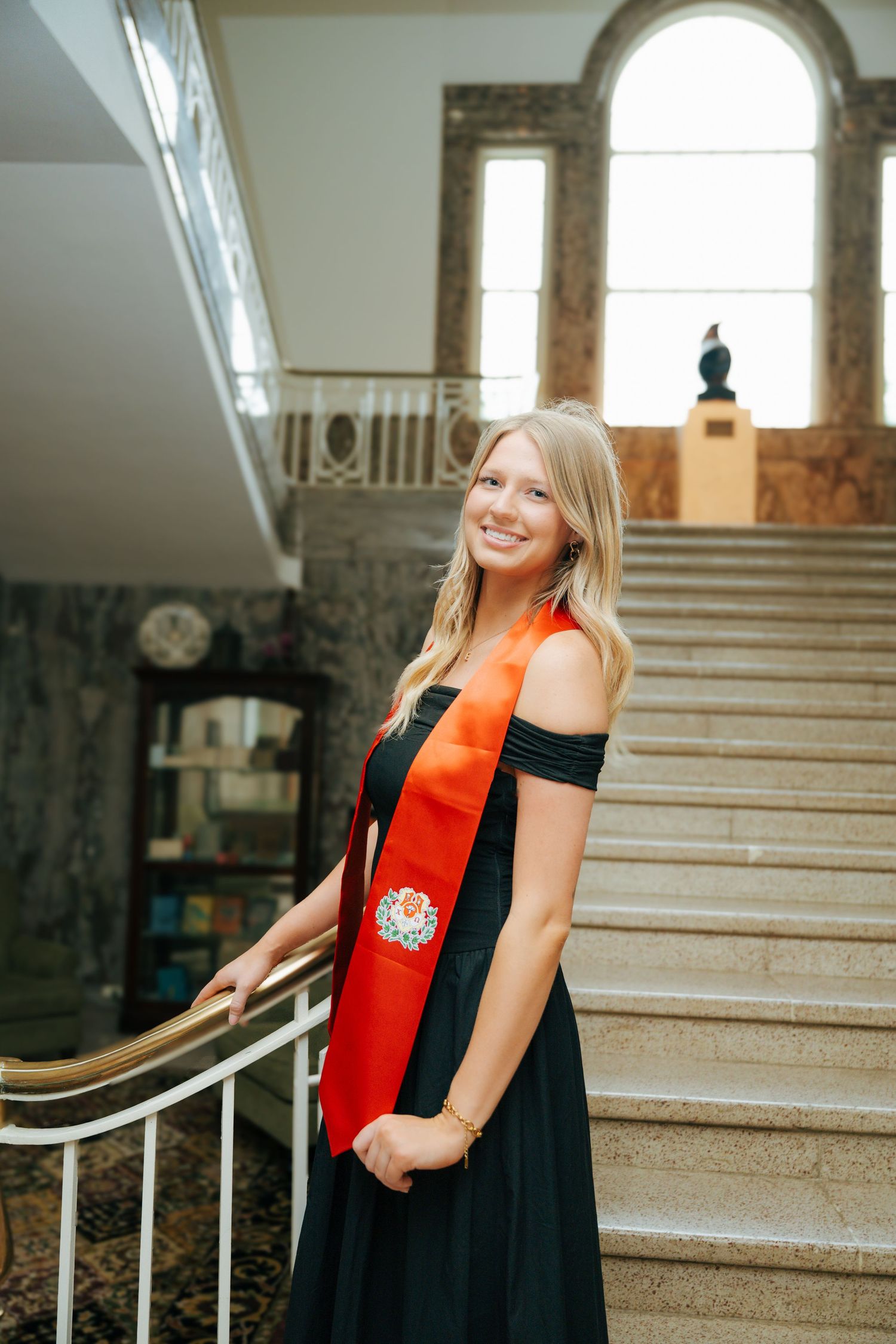 Smiling blonde woman in black gown with red graduation sash poses on marble staircase in elegant building.