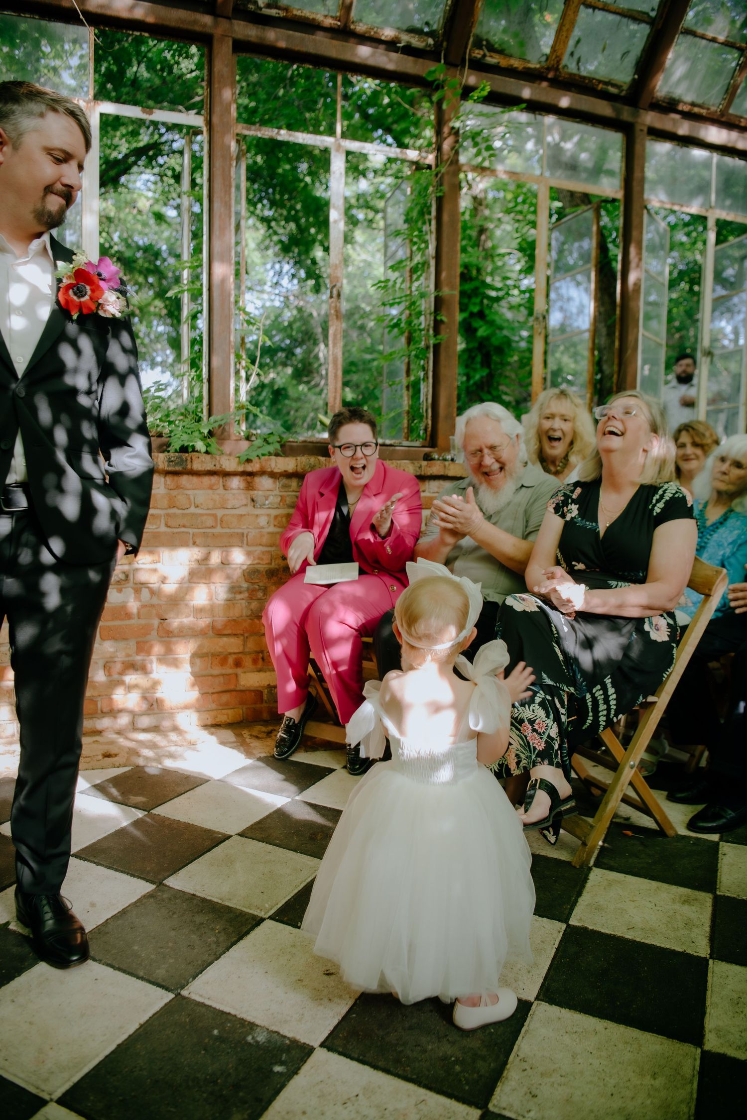 Wedding guests seated in wooden chairs at an indoor venue with checkered flooring.