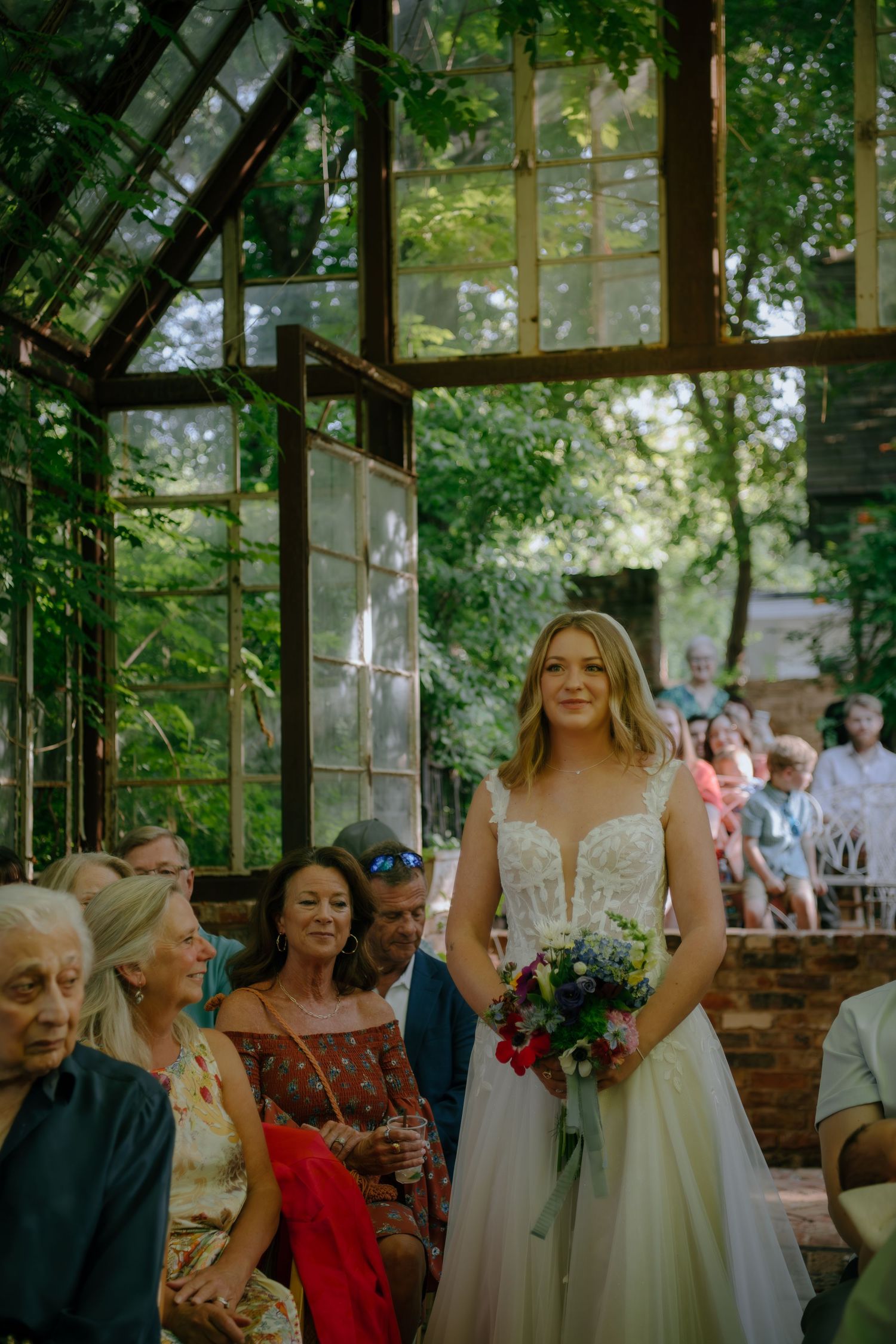 Wedding guests gathered in a sunlit greenhouse venue with brick walls and natural surroundings.