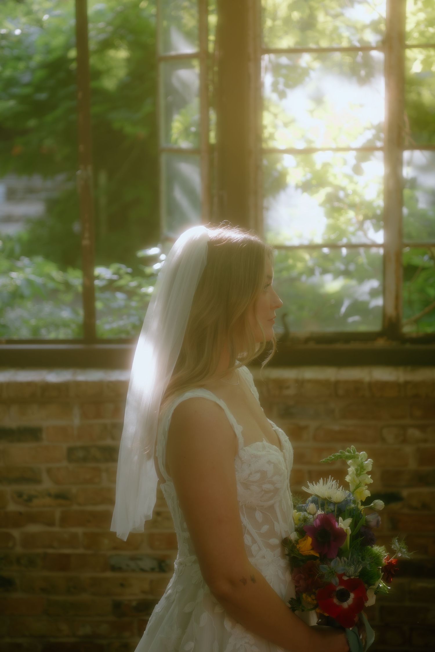 A bride poses with her bouquet by a brick wall and window in soft natural lighting.