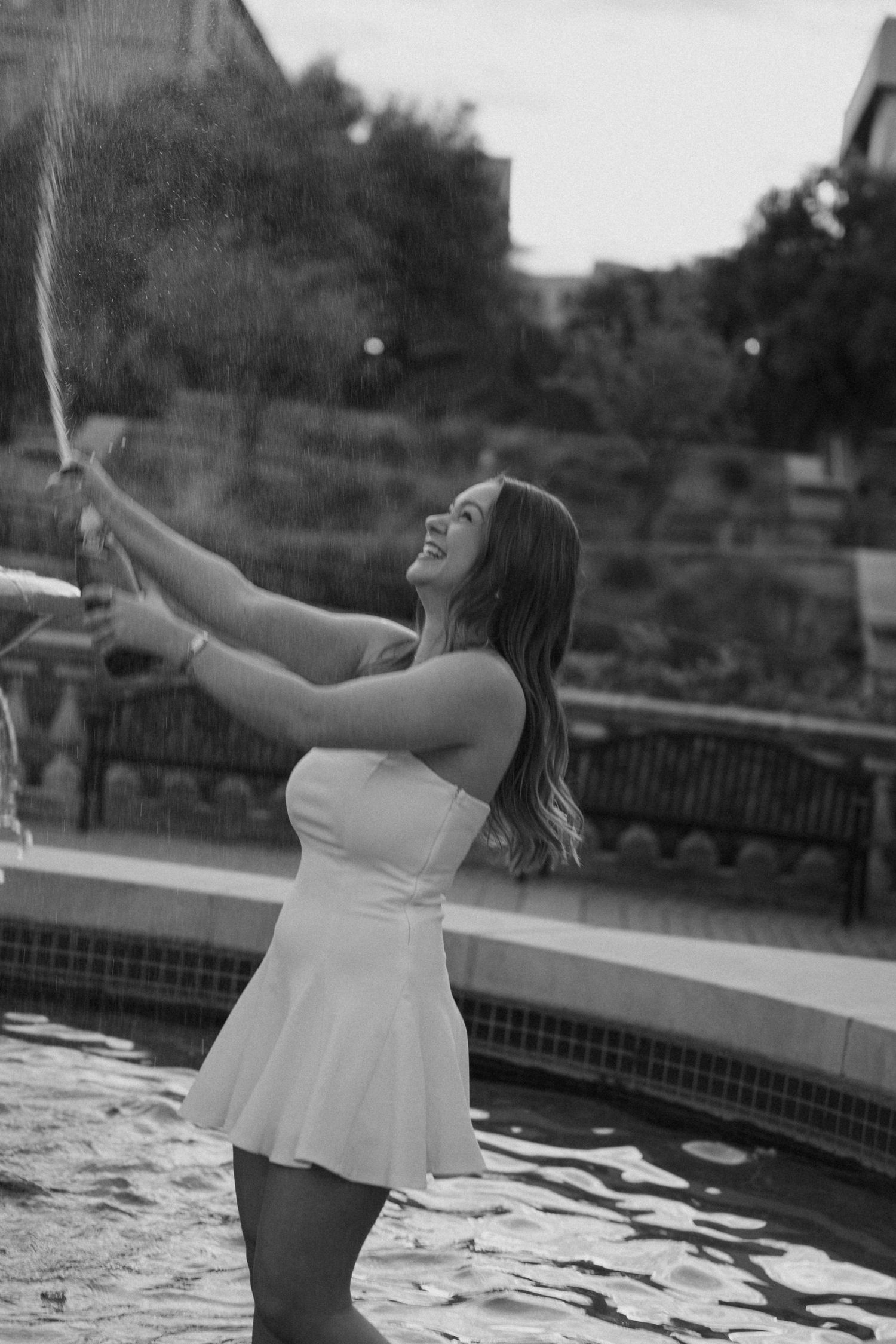 Black and white photo of a smiling woman in a white dress popping champagne by a fountain outdoors.