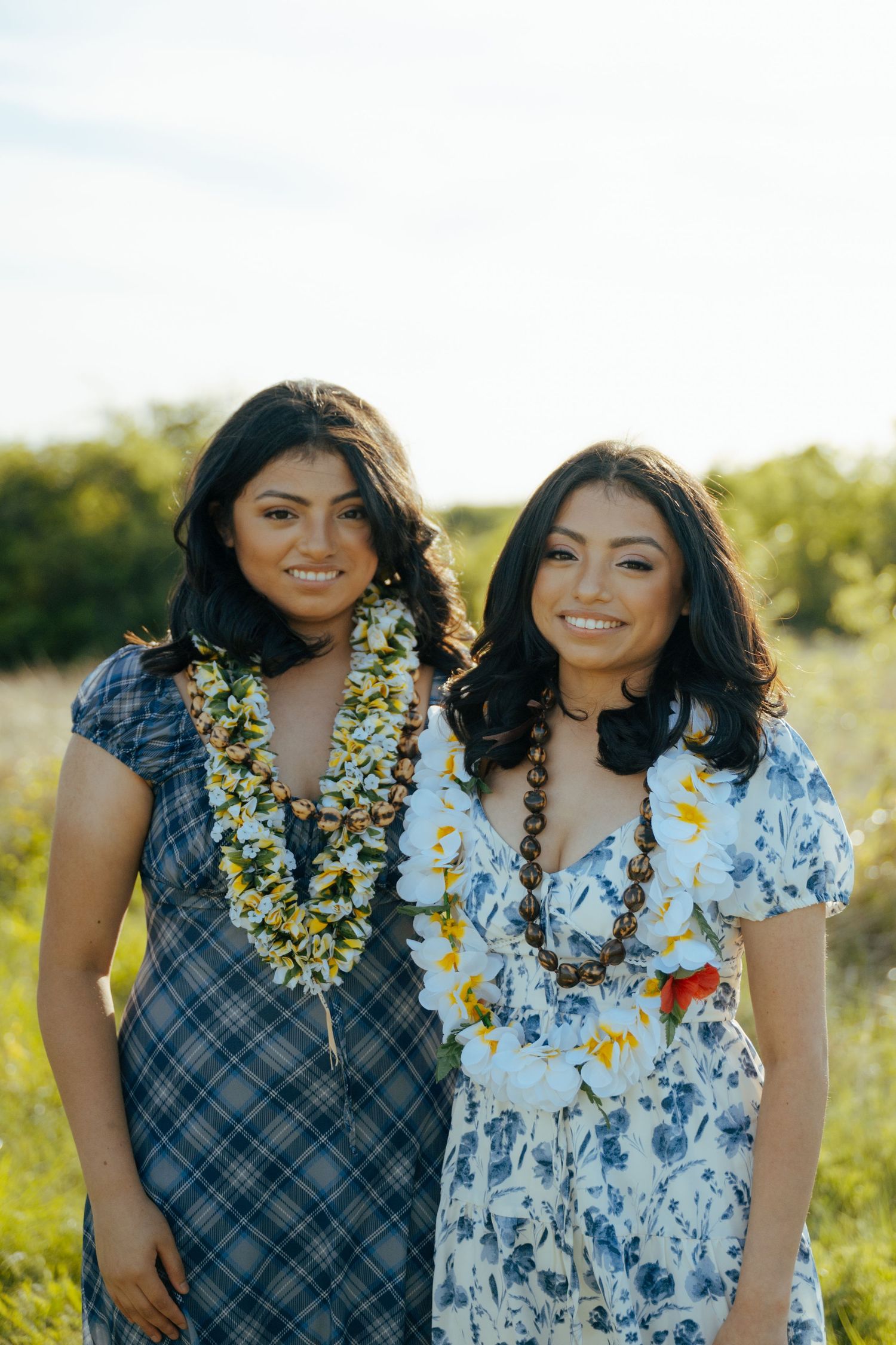 Two smiling young women wearing floral leis and dresses pose outdoors in a sunny green field.