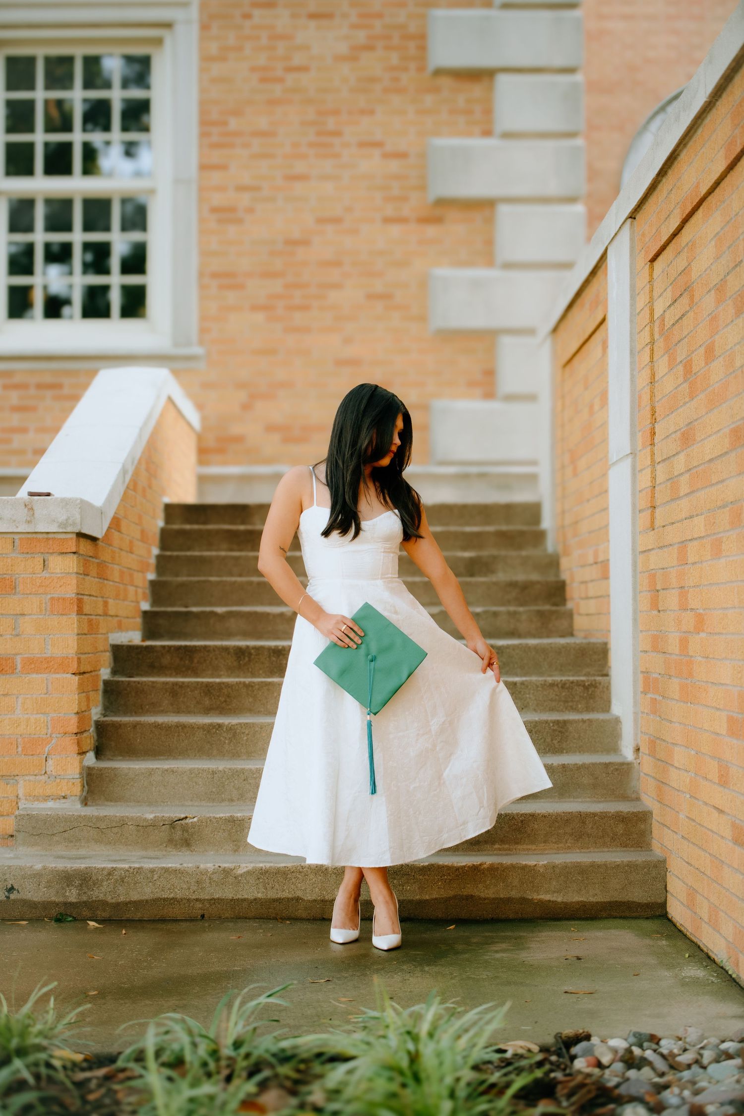 A person in a white dress holds a green book while standing on brick steps near a building.