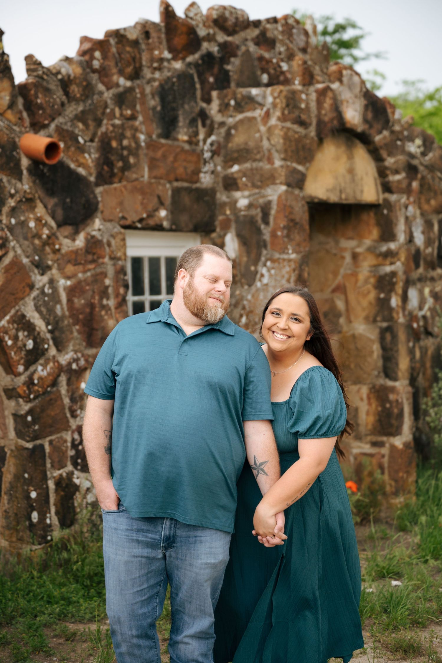 Couple in matching teal outfits holding hands, smiling in front of a rustic stone wall outdoors.