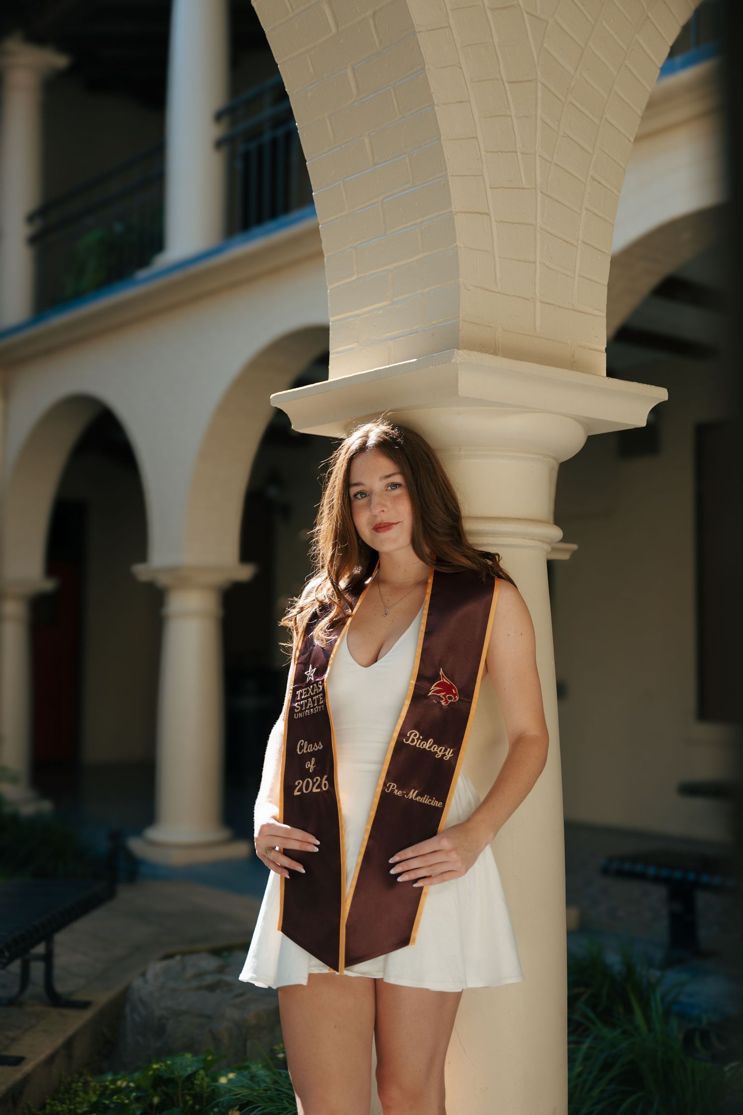 Young woman in white dress wearing maroon graduation stole poses between columns at university campus.