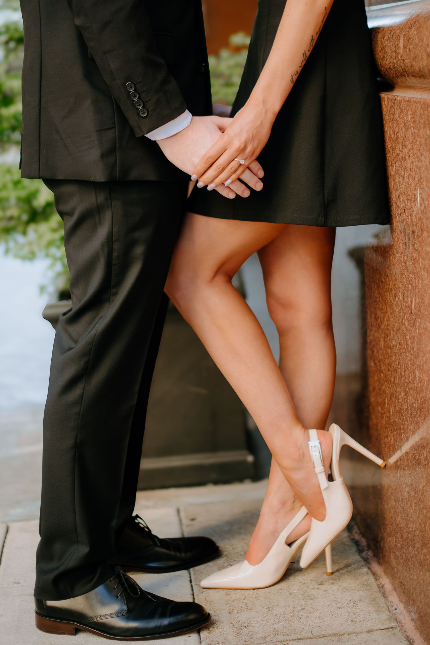 Close-up of couple's feet showing formal dress shoes, with hands clasped together against a stone pillar.